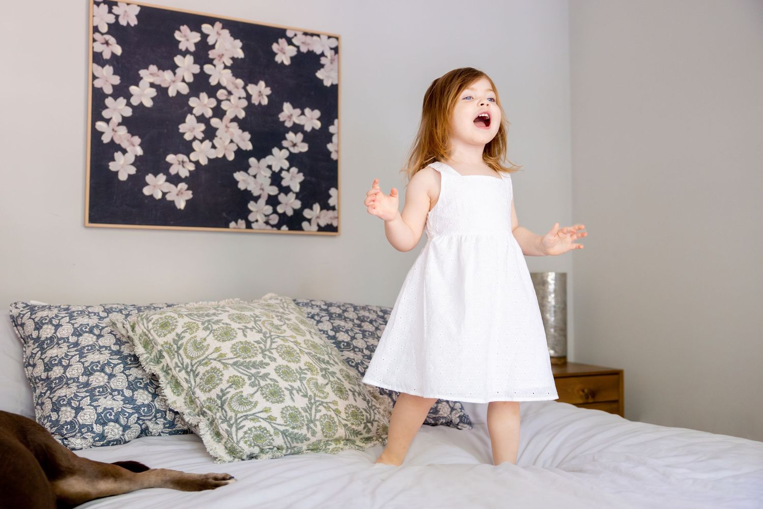 Young child in white dress jumping on bed with floral artwork in background.
