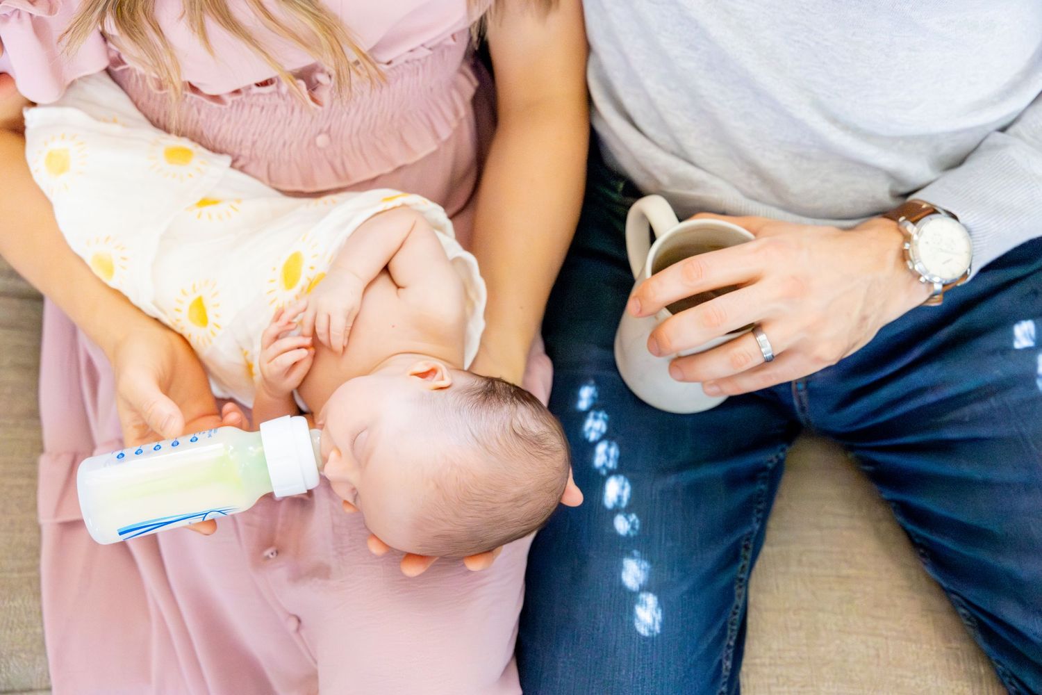 An infant being bottle fed while a person holds a coffee mug on a couch.