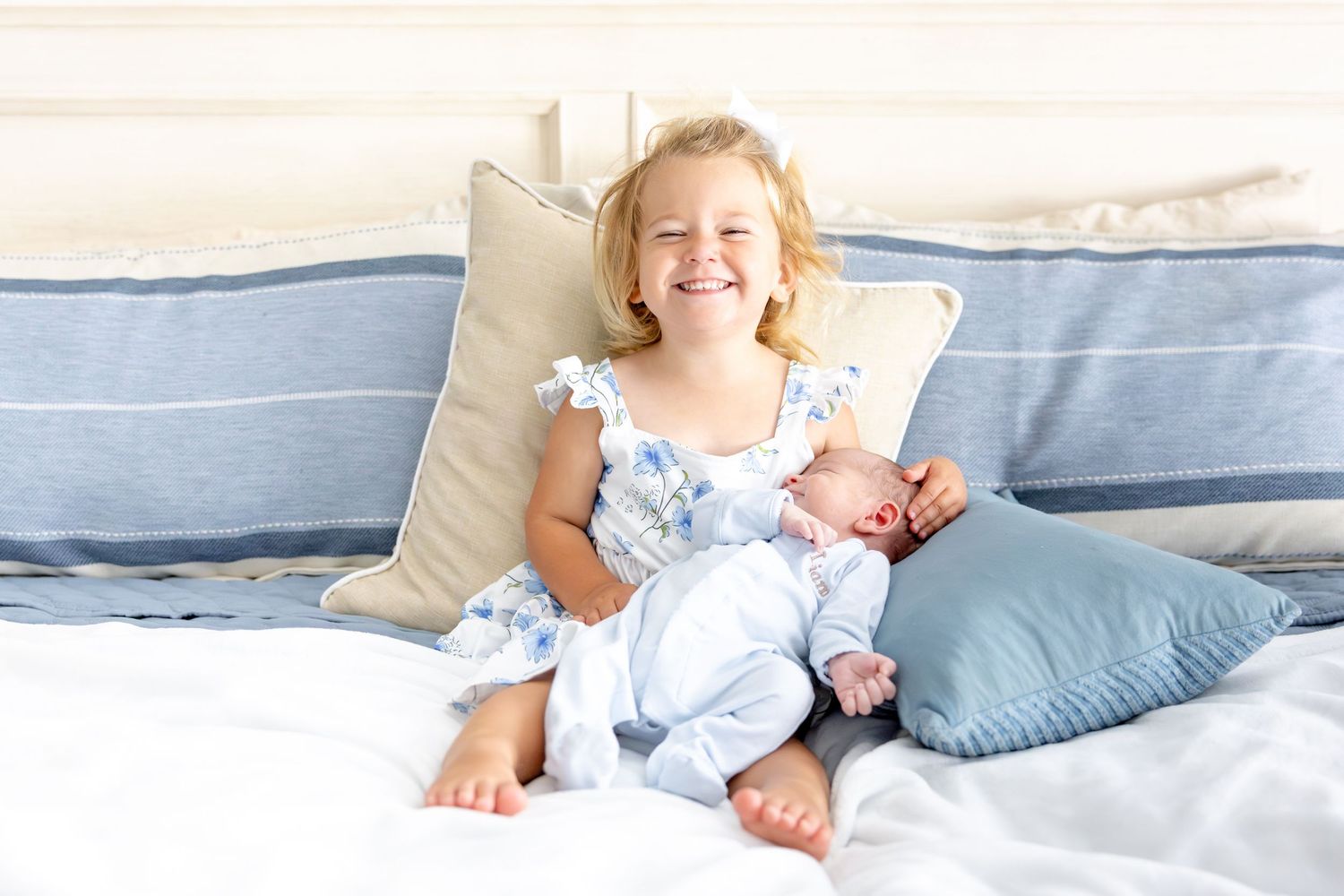 Young child in floral dress sitting happily on bed with blue pillows.