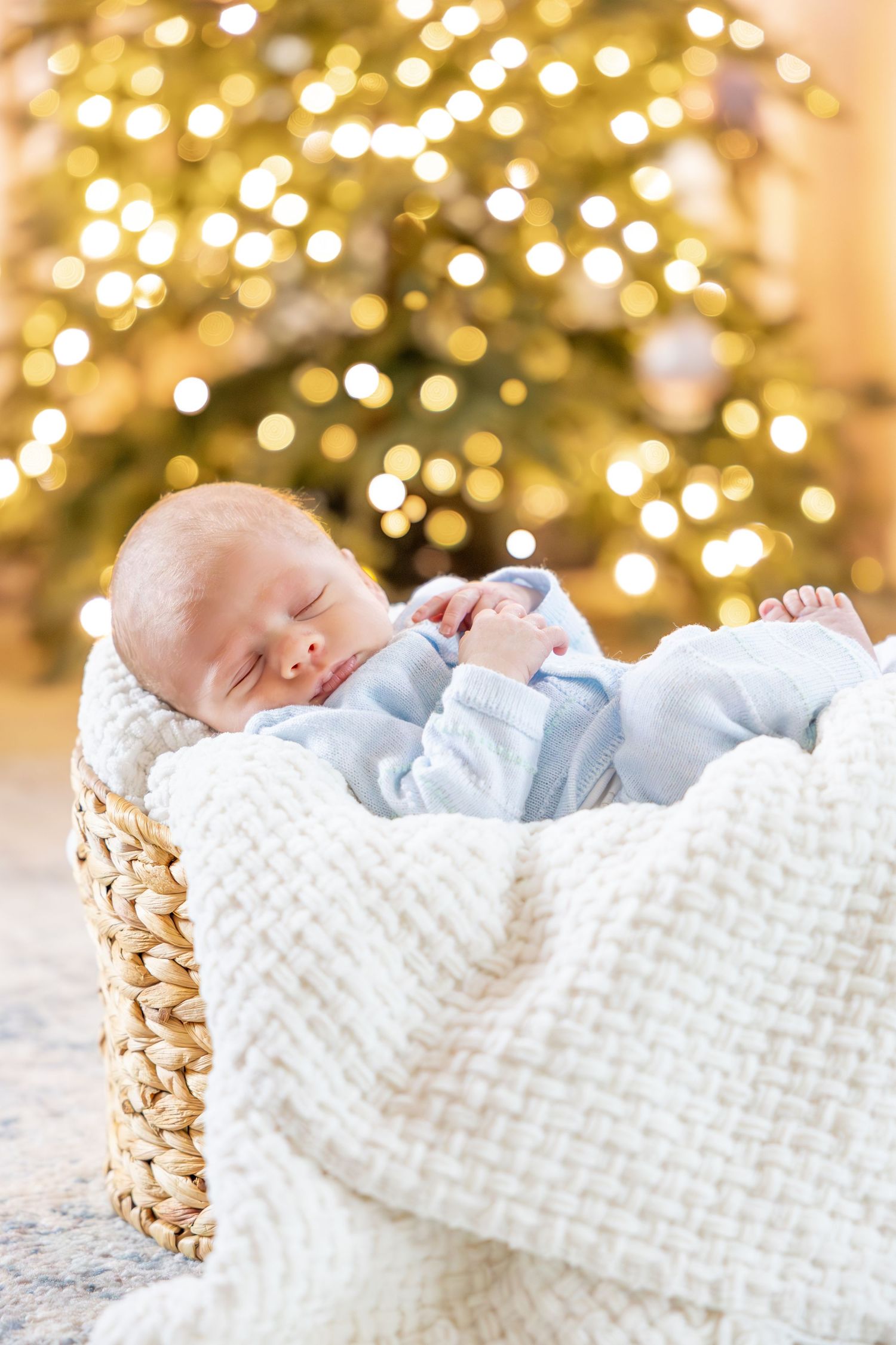 Sleeping infant in white blanket resting in woven basket with Christmas lights behind.