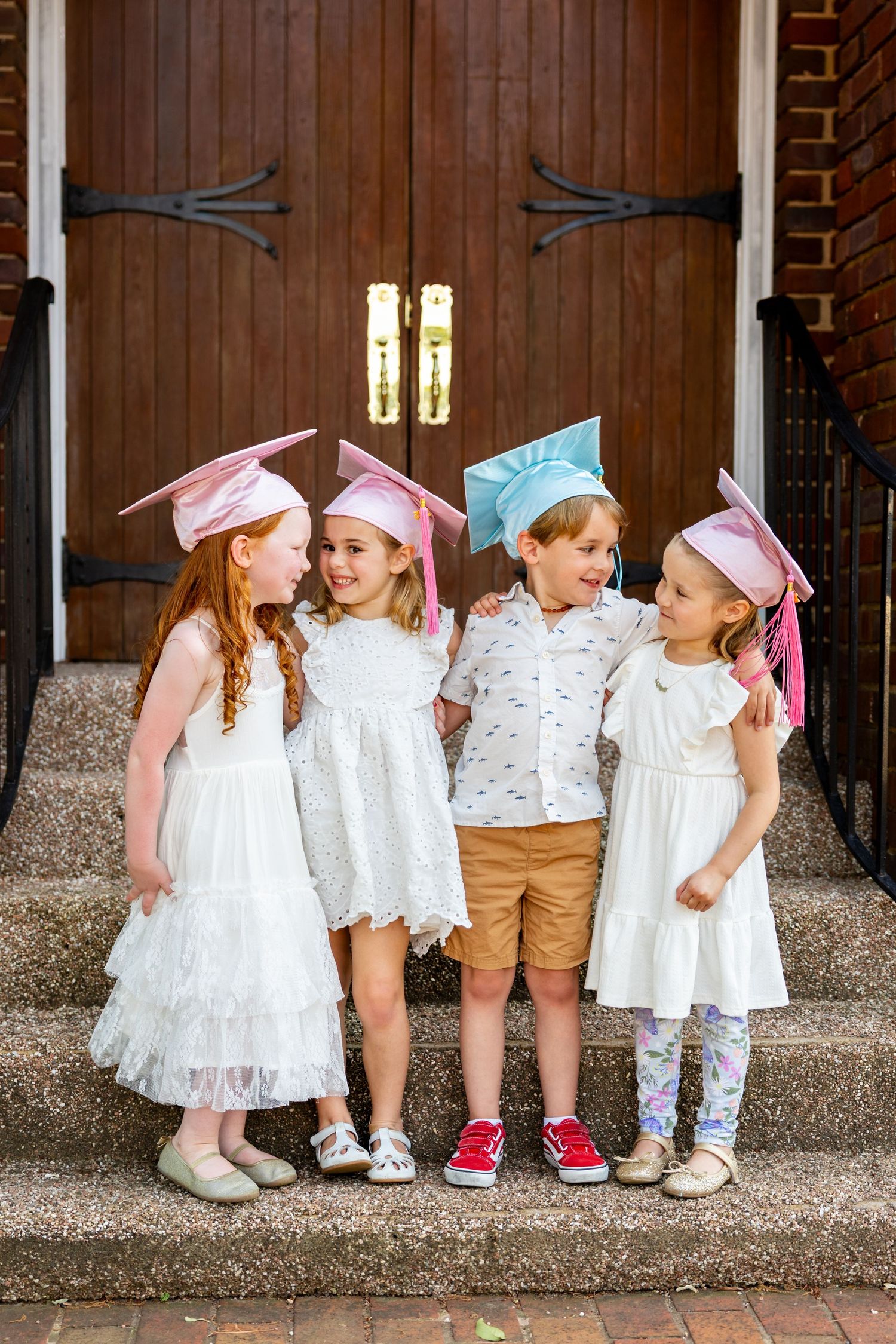 Group of small preschool children in white outfits and pink and blue graduation caps standing together against a brick wall.