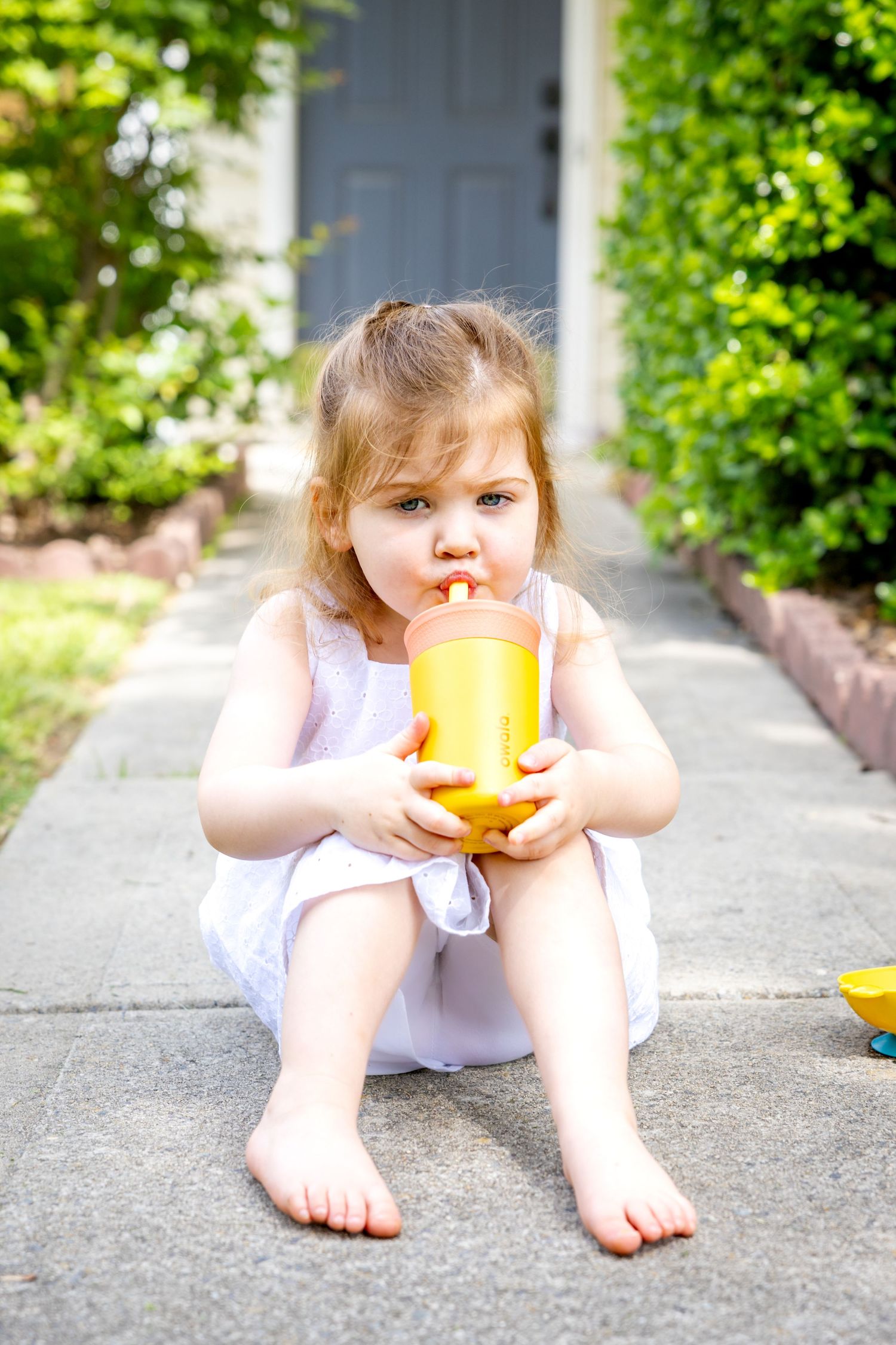 Small child in white dress sitting on concrete path drinking from yellow cup.