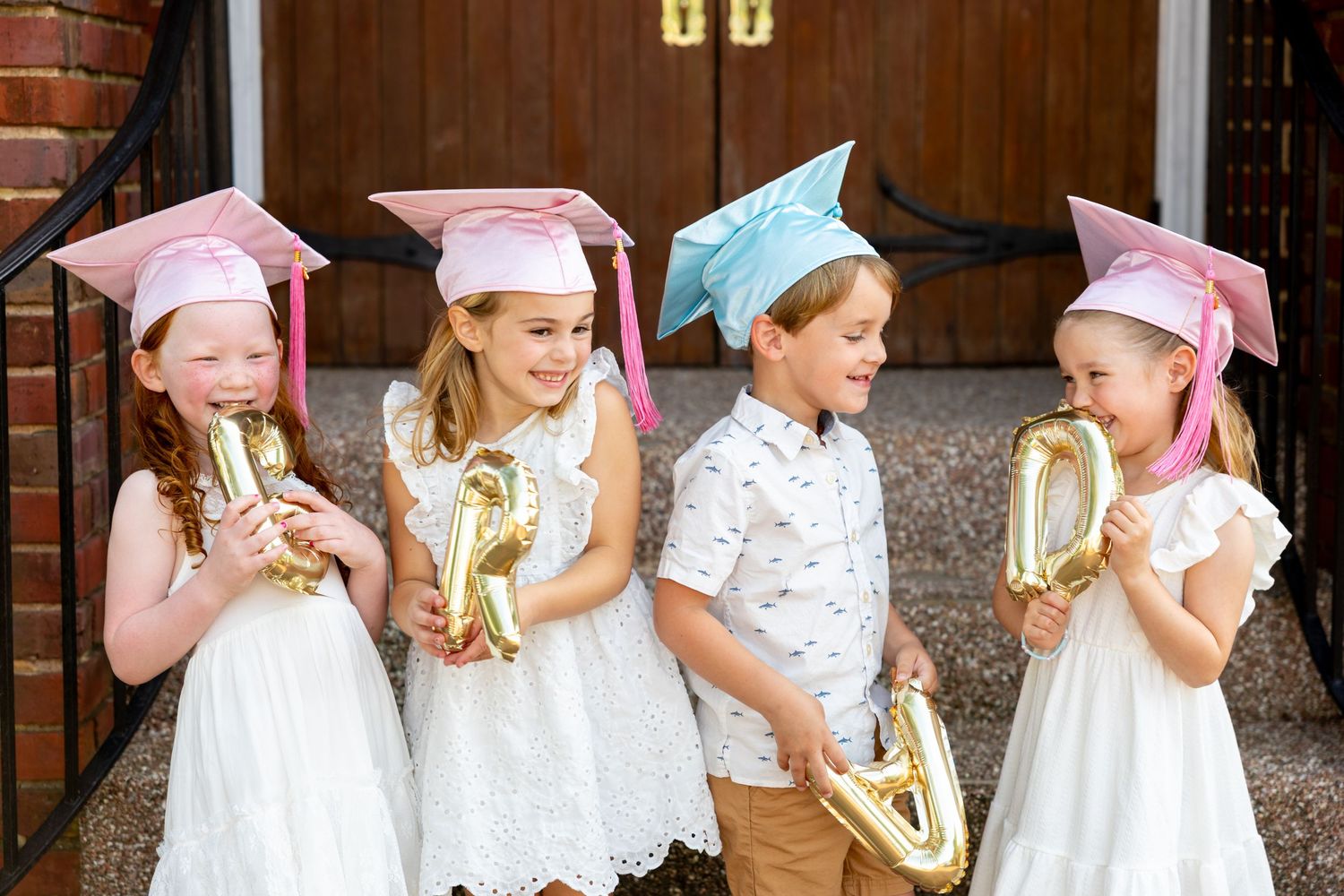 Preschool graduates in white dresses with colorful caps stand together against brick backdrop.