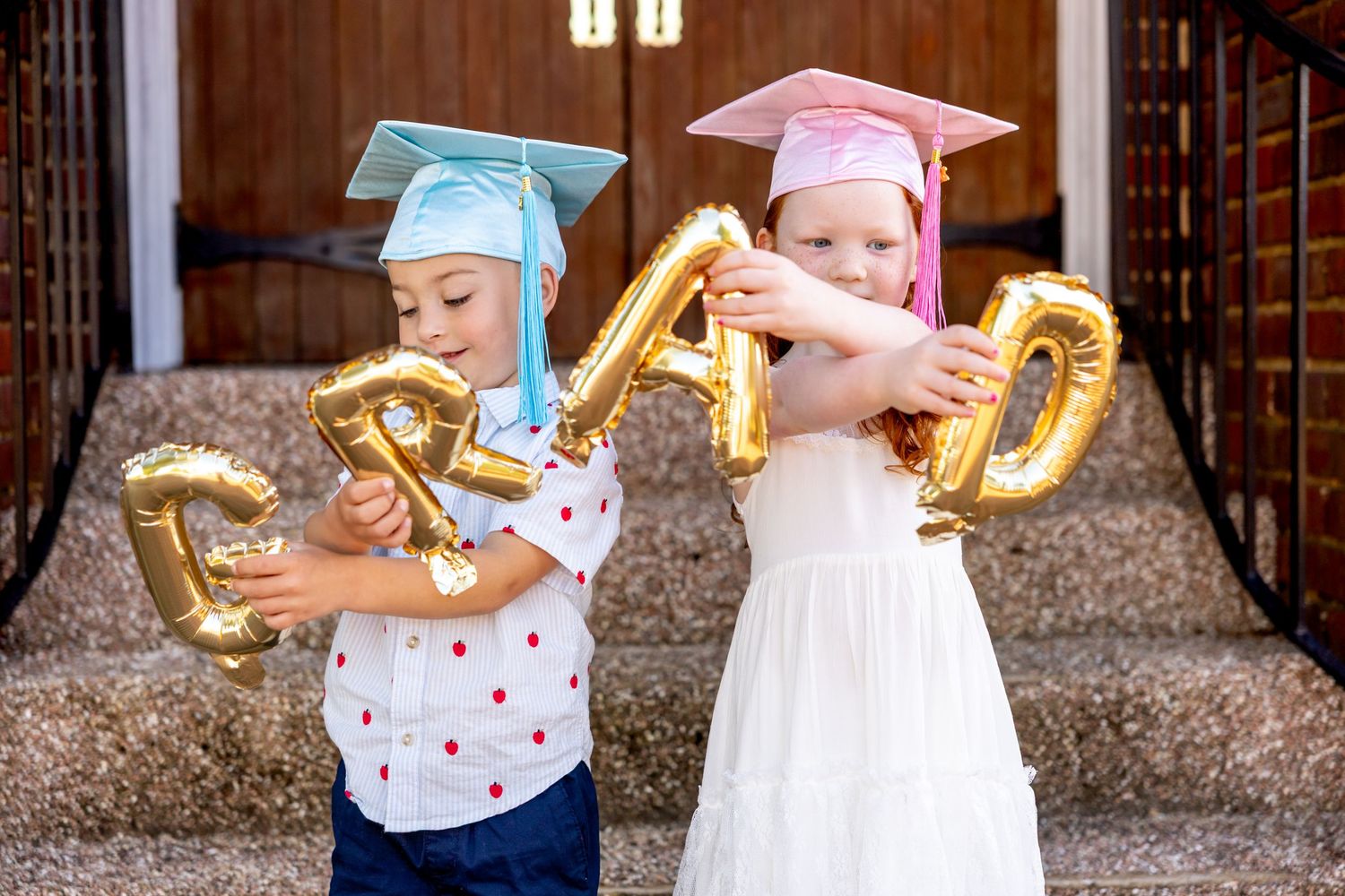 Two young preschool graduates holding gold number balloons.