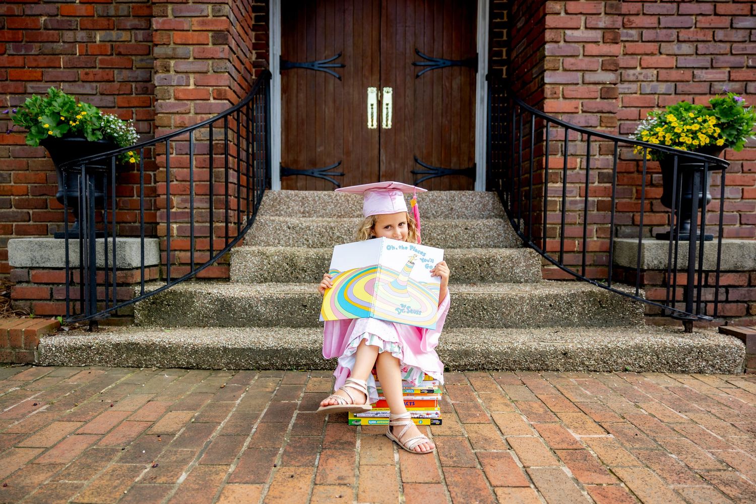 Wide angle view of brick steps and wooden door with preschool graduate reading a book.