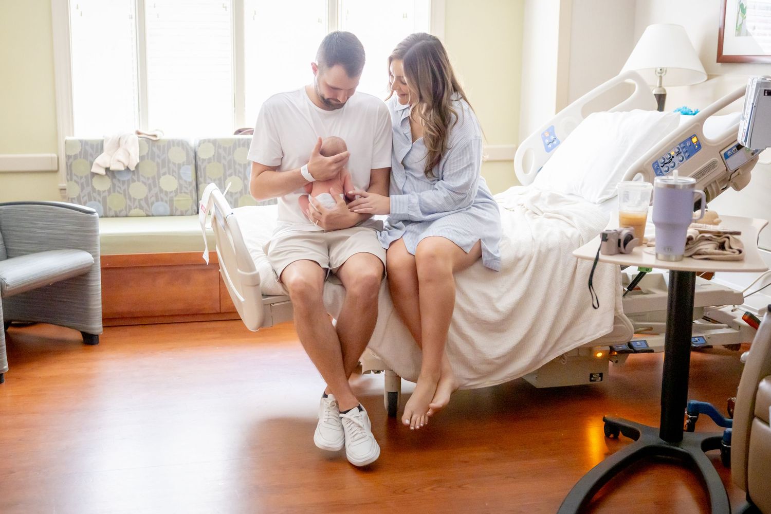 New parents sitting on hospital bed with their newborn in bright room.