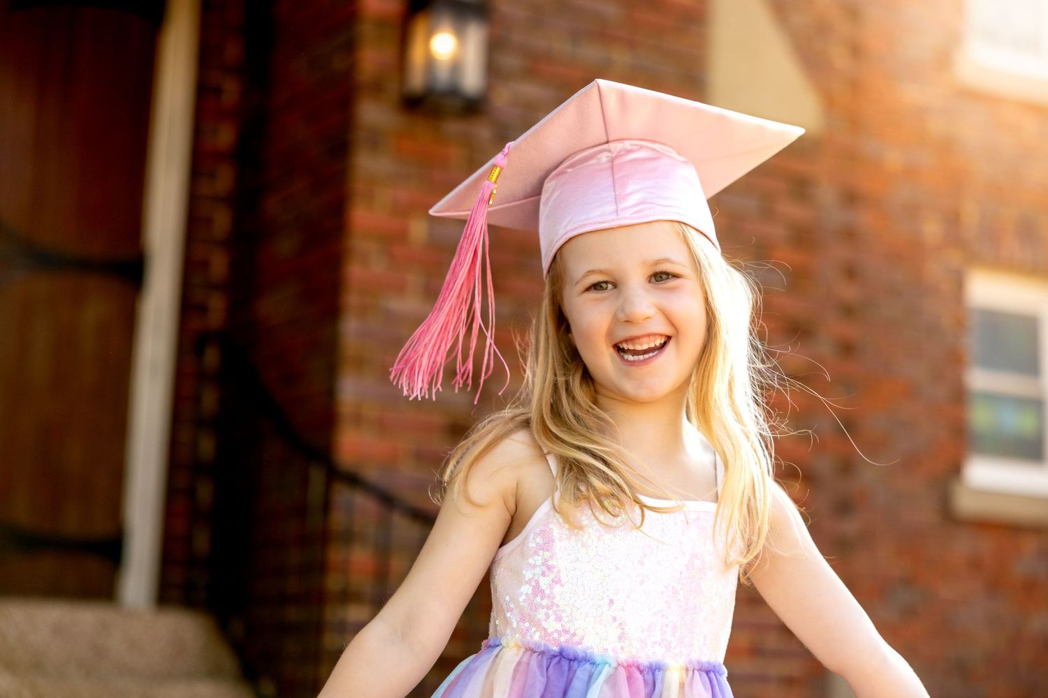 Preschool graduate in sparkly dress and pink cap poses joyfully in golden sunlight.