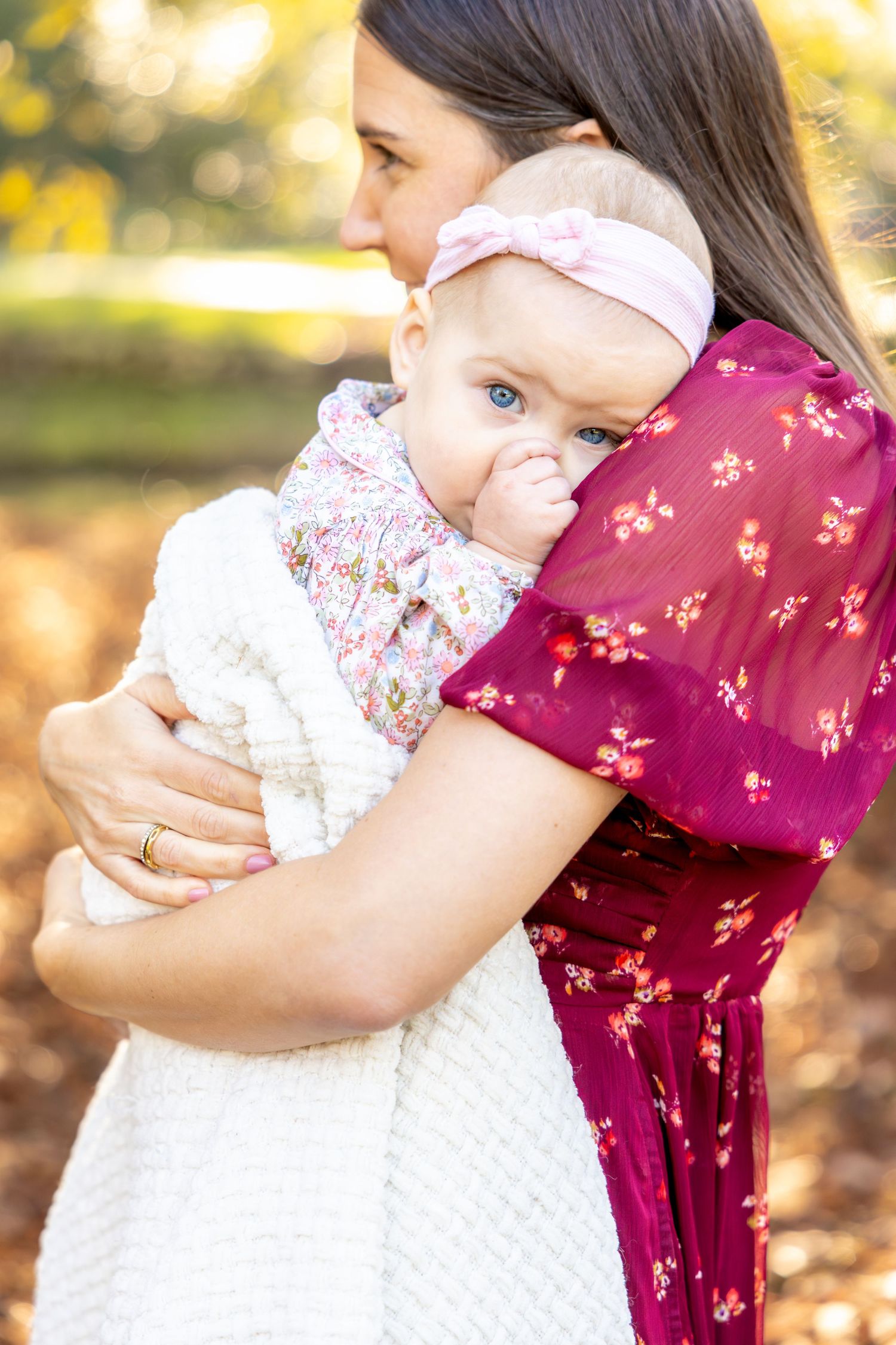 Person in burgundy floral dress holding baby in white lace dress outdoors at Glen Cairn Gardens in Rock Hill, SC.