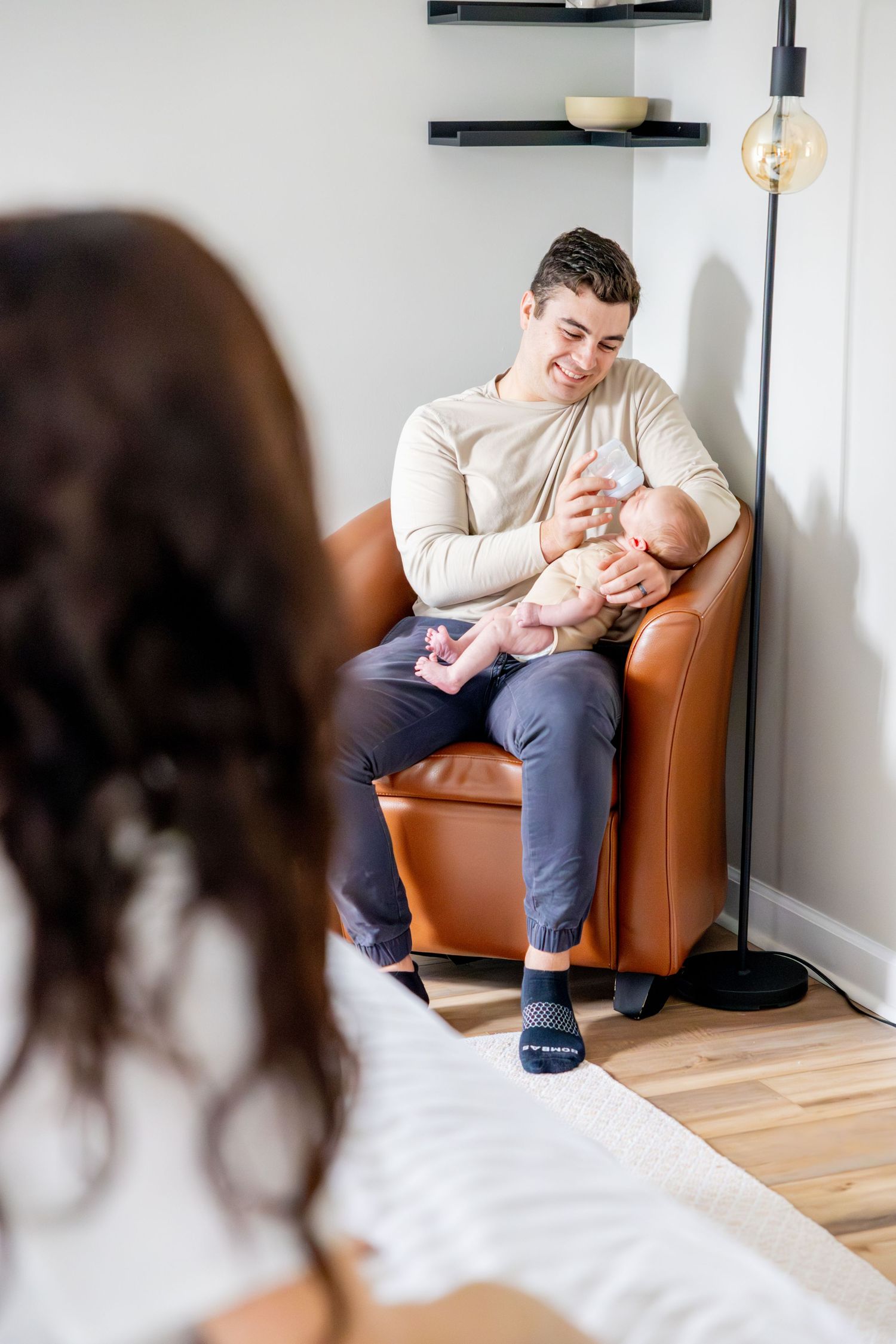 Parent sits in leather chair holding and feeding newborn baby while being photographed in minimalist modern living room.