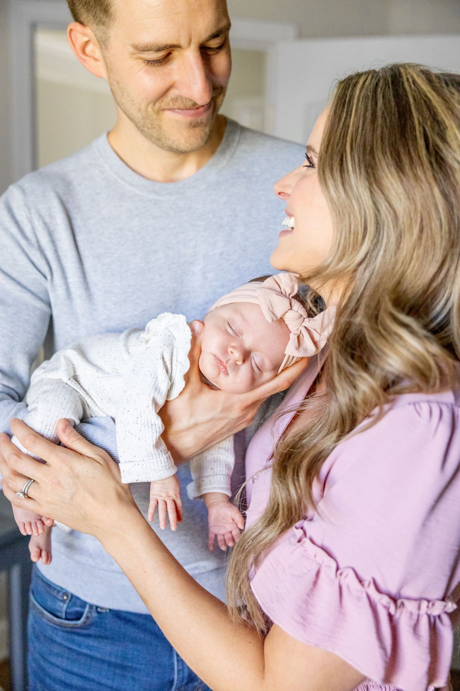 Family photoshoot captures sweet moments between parents and their sleeping newborn in matching grey and pink outfits.