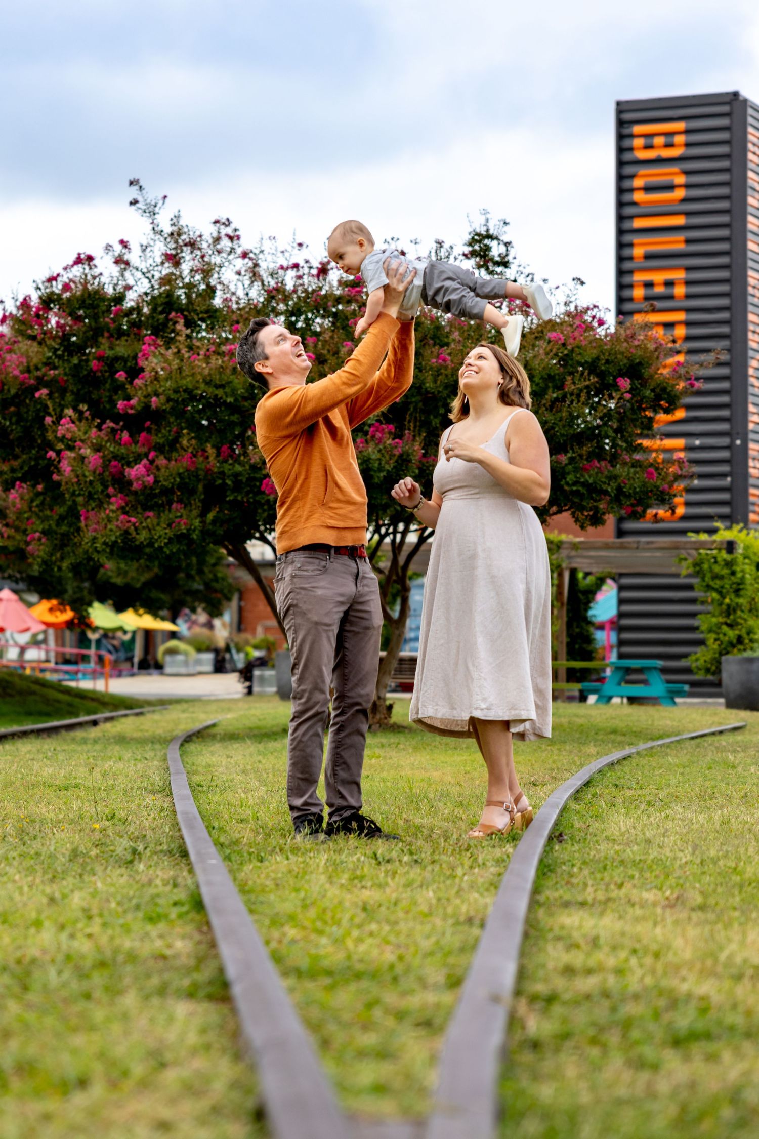 Couple shares joyful moments together on grassy lawn with tall building in background at Camp North End in Charlotte, NC.