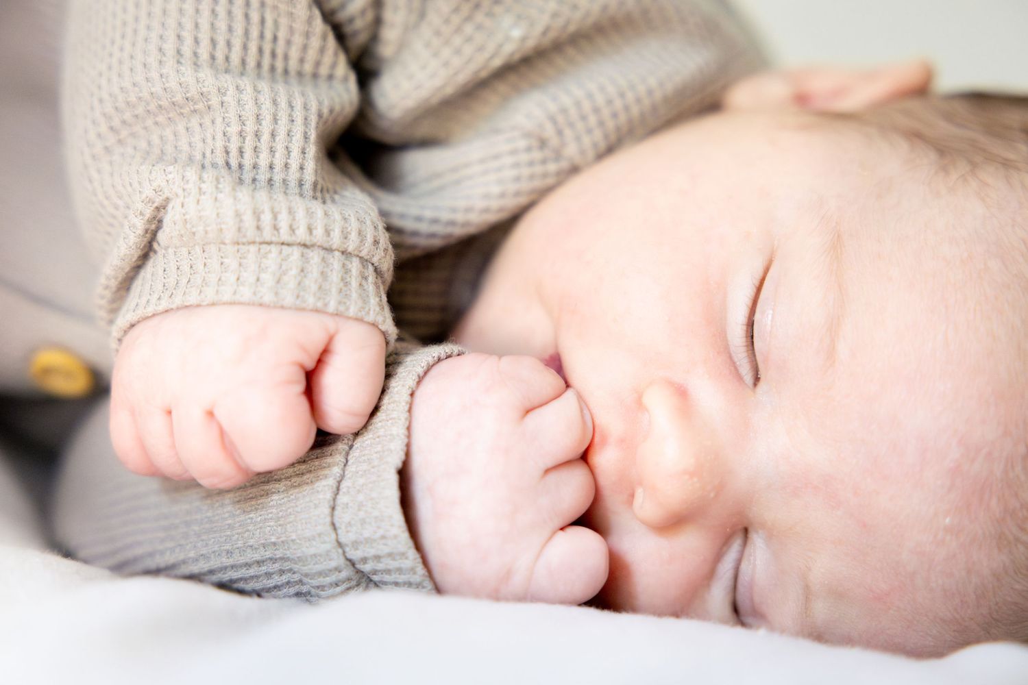 Sleeping infant in gray sweater with hands near face on white bedding.