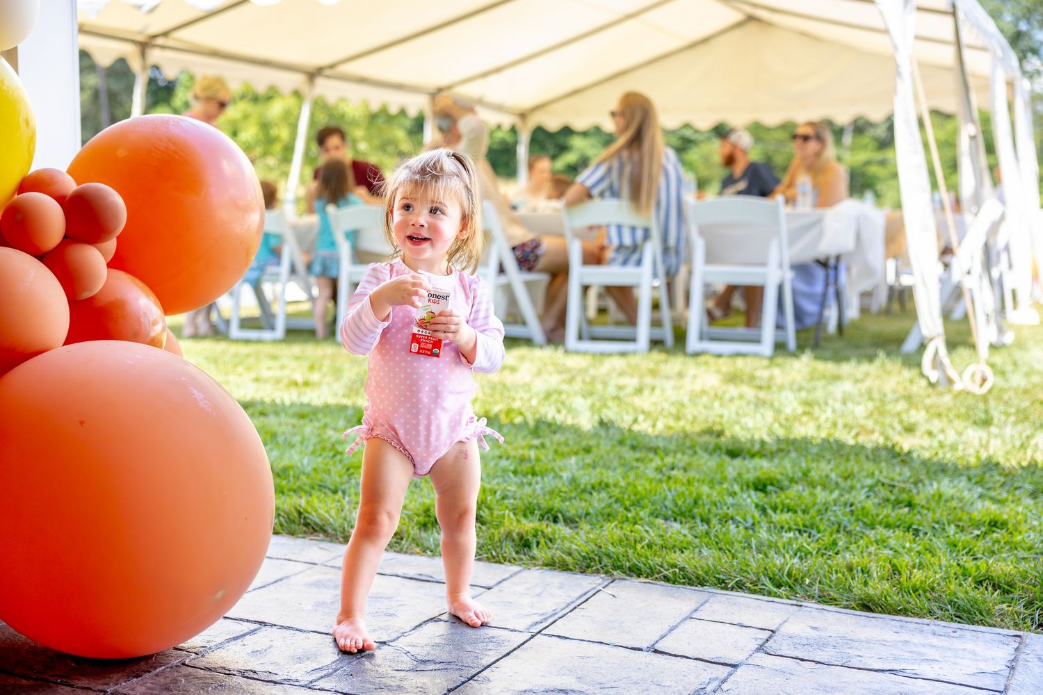 Outdoor birthday celebration with toddler walking among orange and yellow balloons.