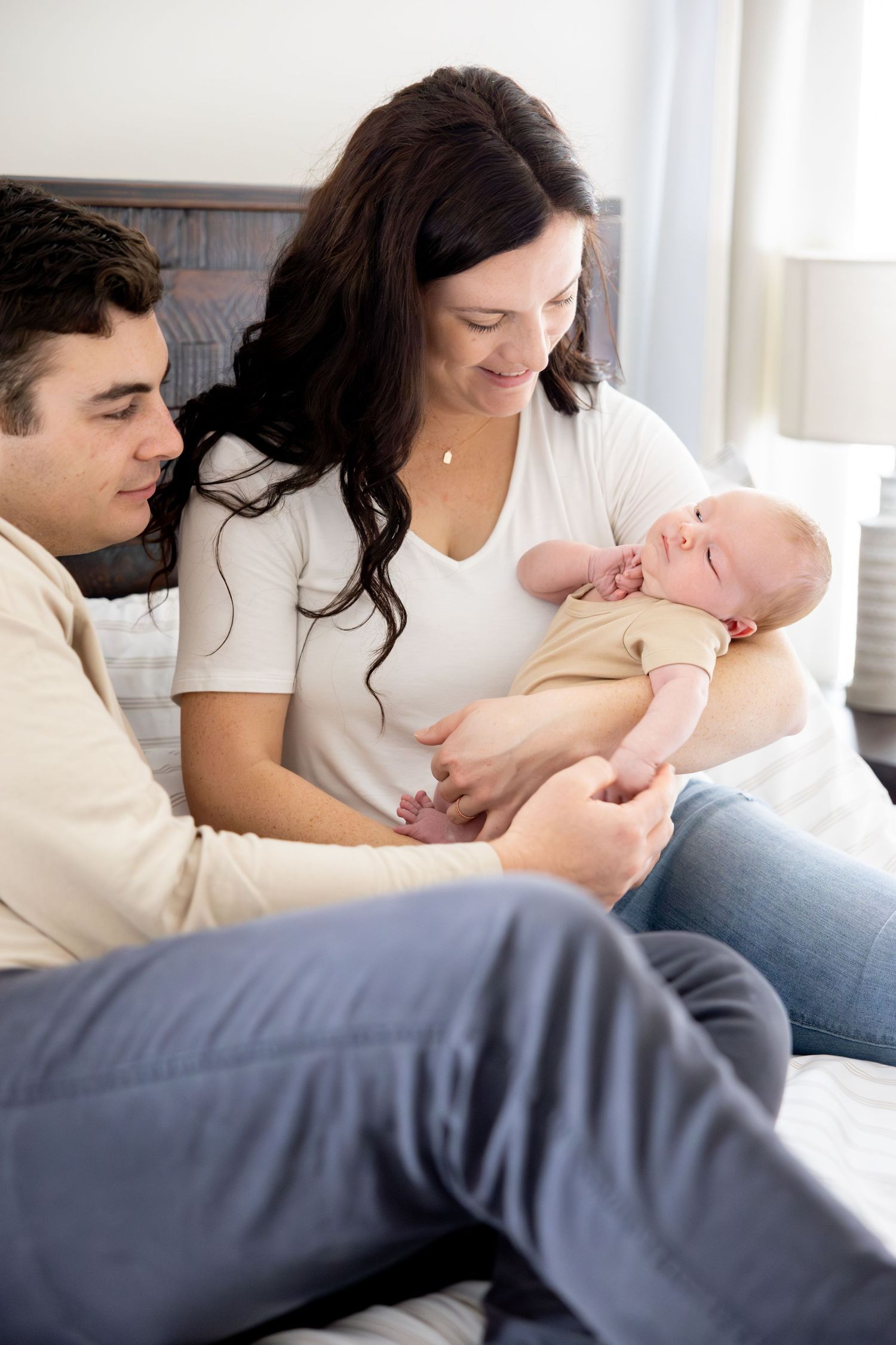 Parents sitting on bed sharing tender moment with newborn baby in casual lifestyle portrait.