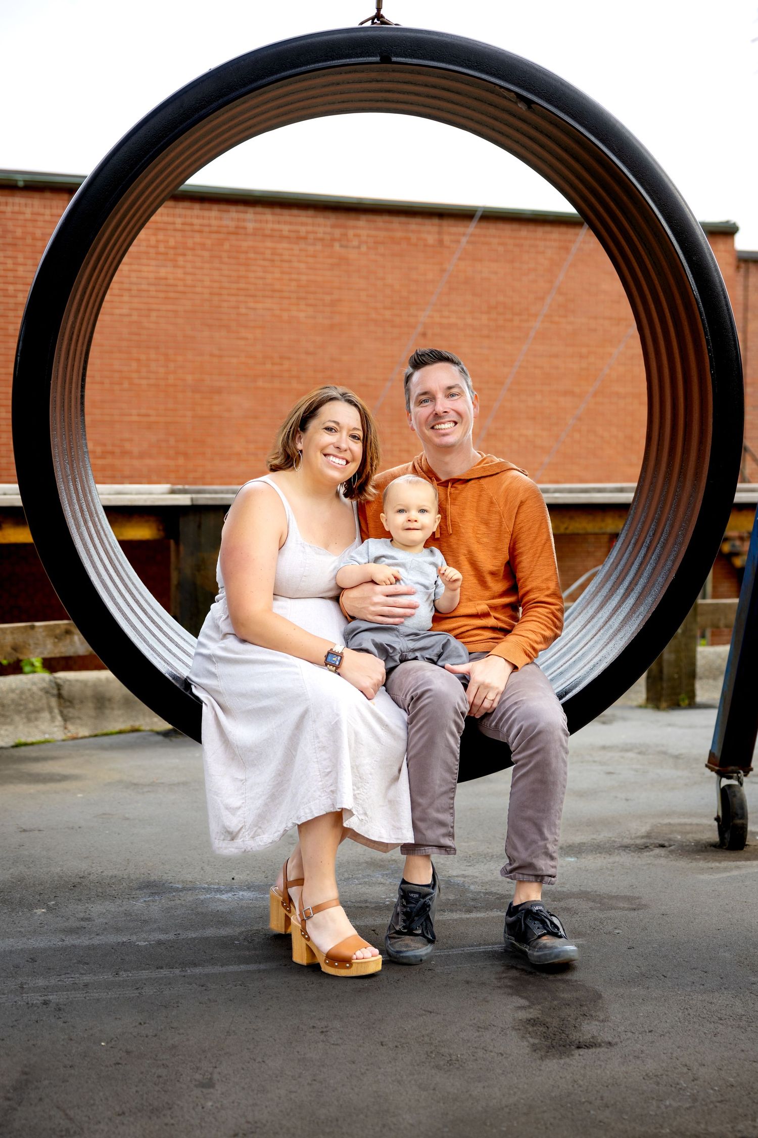 Family sitting together inside large metal ring structure against brick wall at Camp North End in Charlotte, NC.