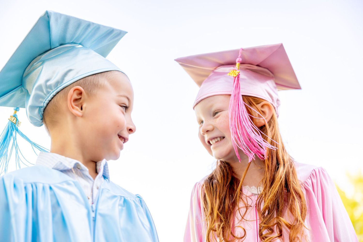 Two preschool graduates in pink and blue caps and gowns standing together.