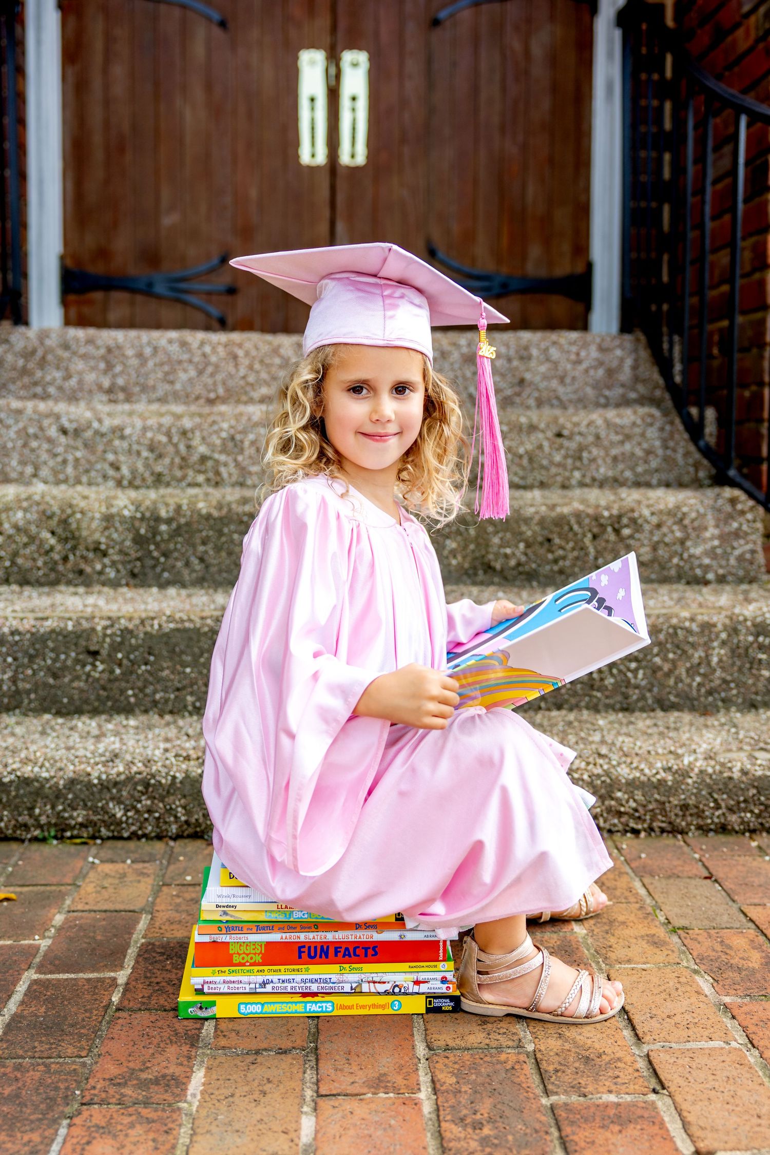 Child in pink preschool graduation cap sits on pile of books in front of concrete steps.
