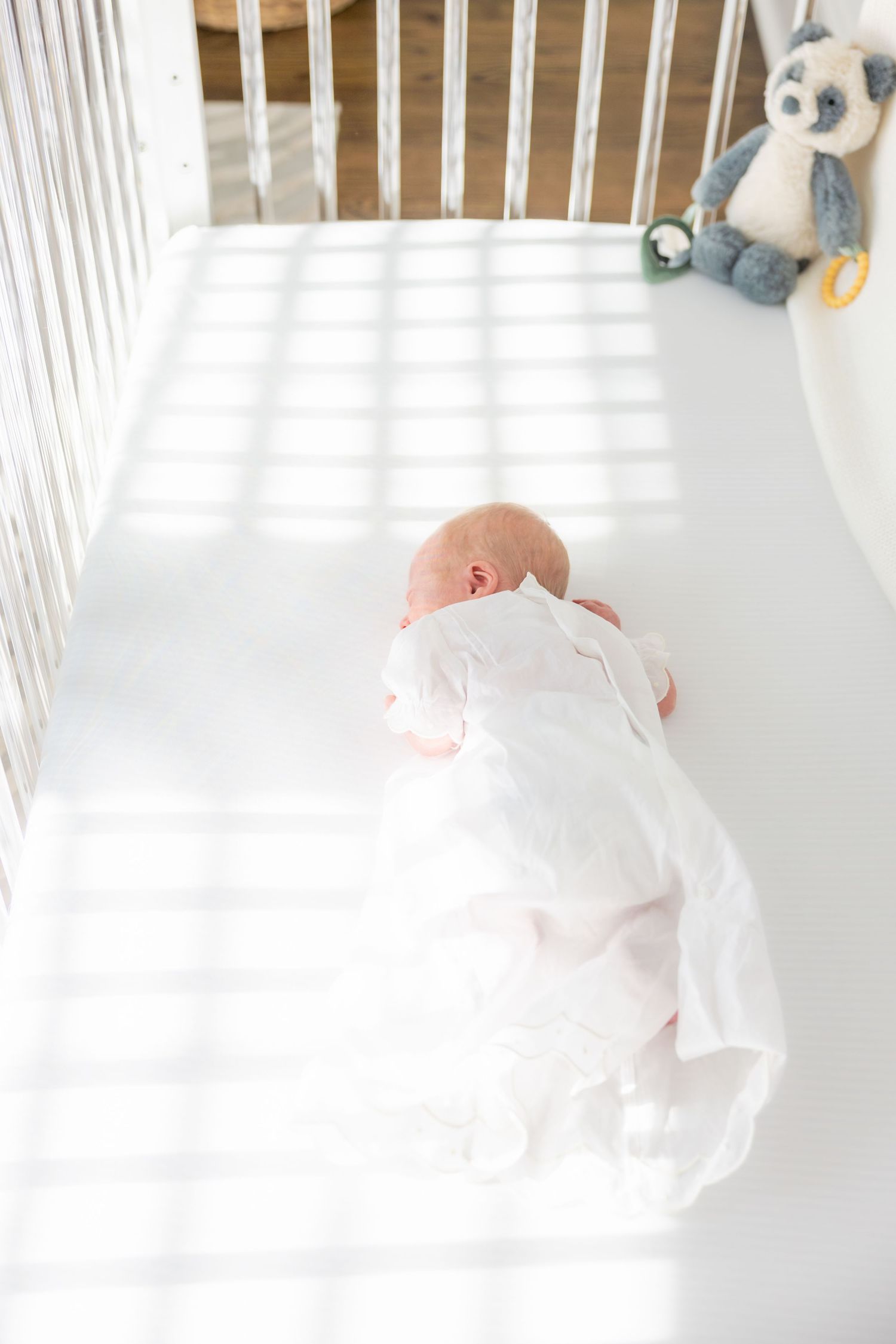 Sleeping baby in white clothing rests peacefully in a white crib with stuffed animal nearby.