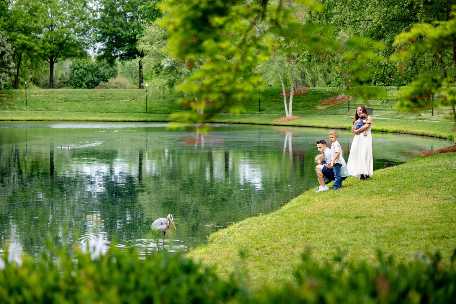 Family relaxing by a serene pond surrounded by lush greenery in a tranquil park setting.