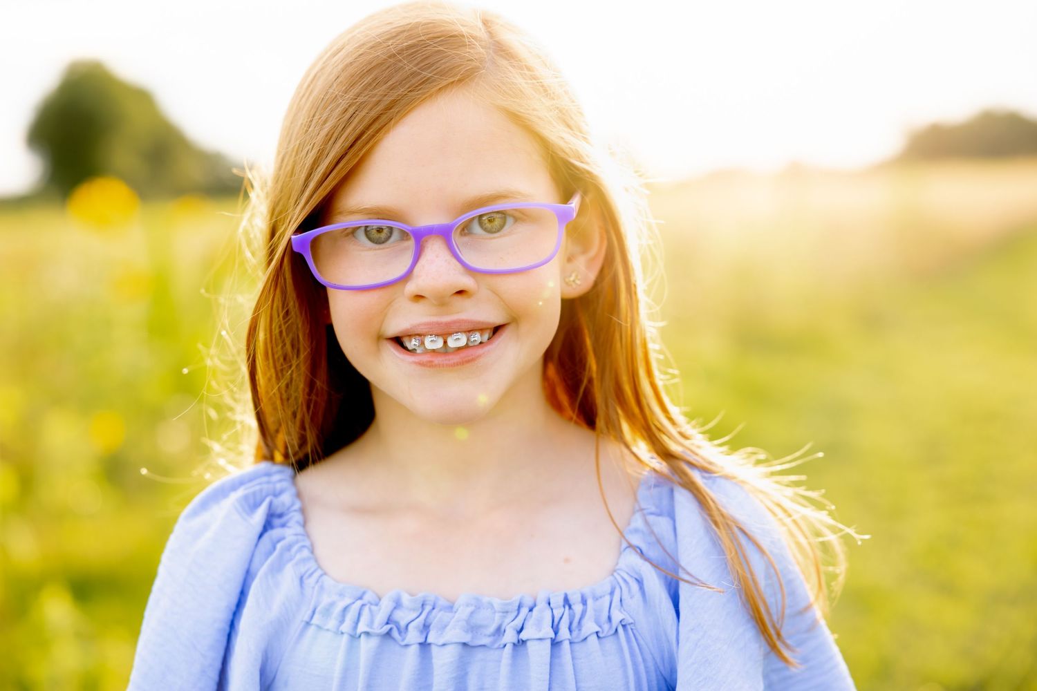 Bright smile and purple glasses against golden sunset in meadow wearing blue ruffled dress at Blue Hill Garden in Monroe, NC.
