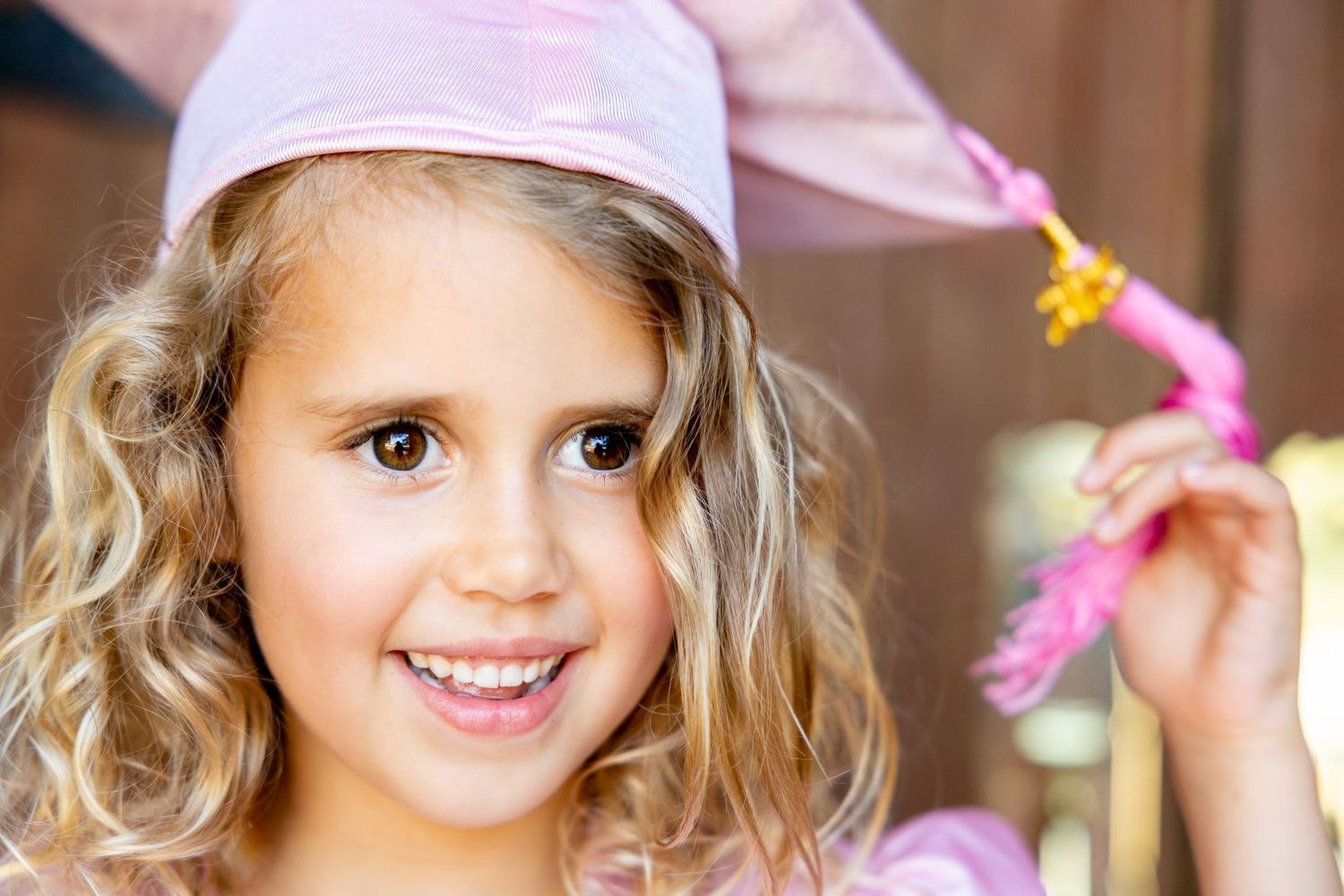 Young preschool graduation child in pink cap smiling against wooden background.