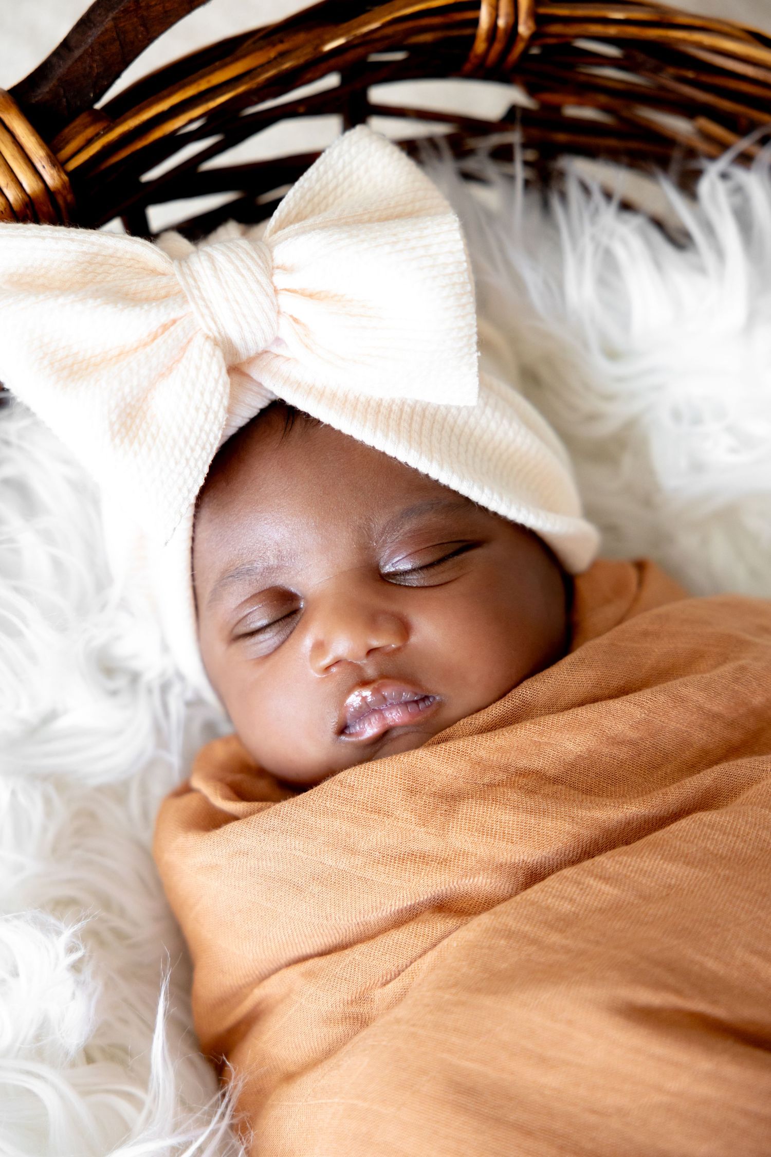 Peaceful sleeping baby wrapped in orange fabric resting in woven basket with white fur lining.