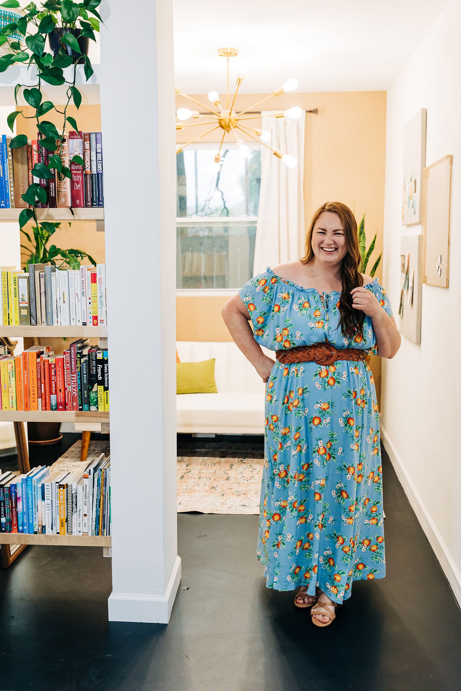 Woman in floral maxi dress poses in modern home interior with bookshelf.