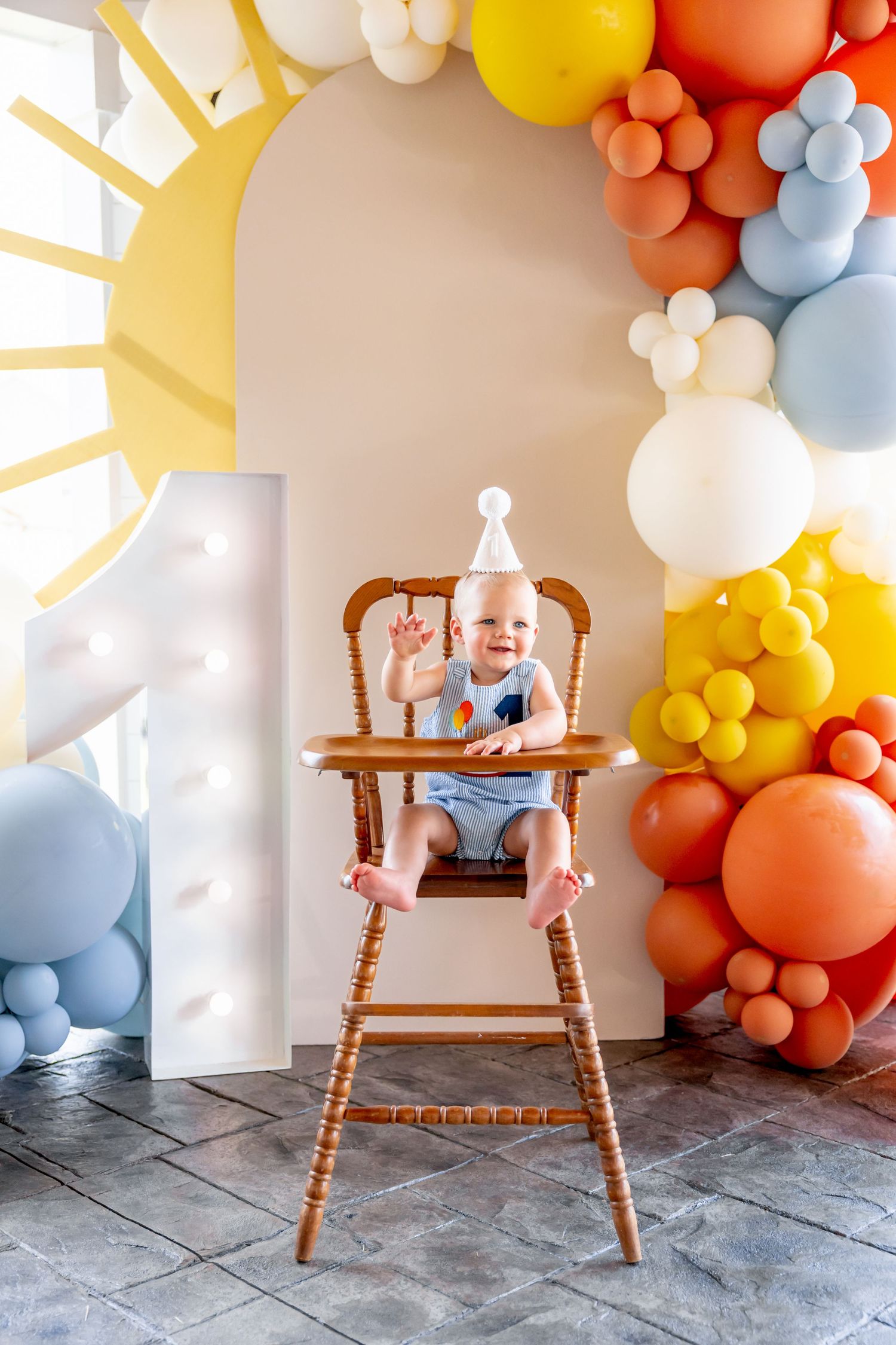 Baby milestone photos with colorful balloon arch and wooden high chair decorations.