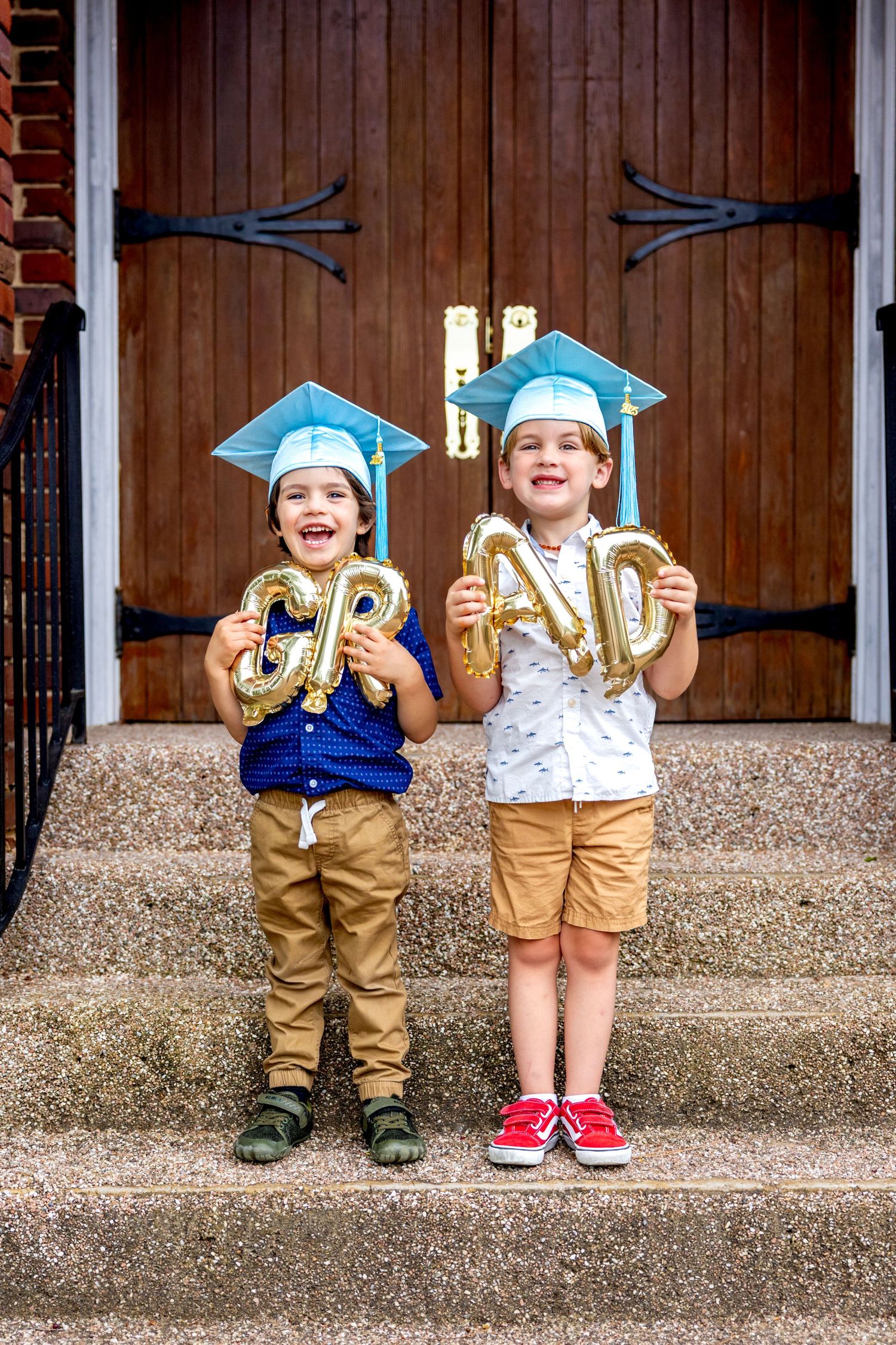 Two preschool graduates in blue caps and khaki outfits standing on concrete steps with gold balloons.