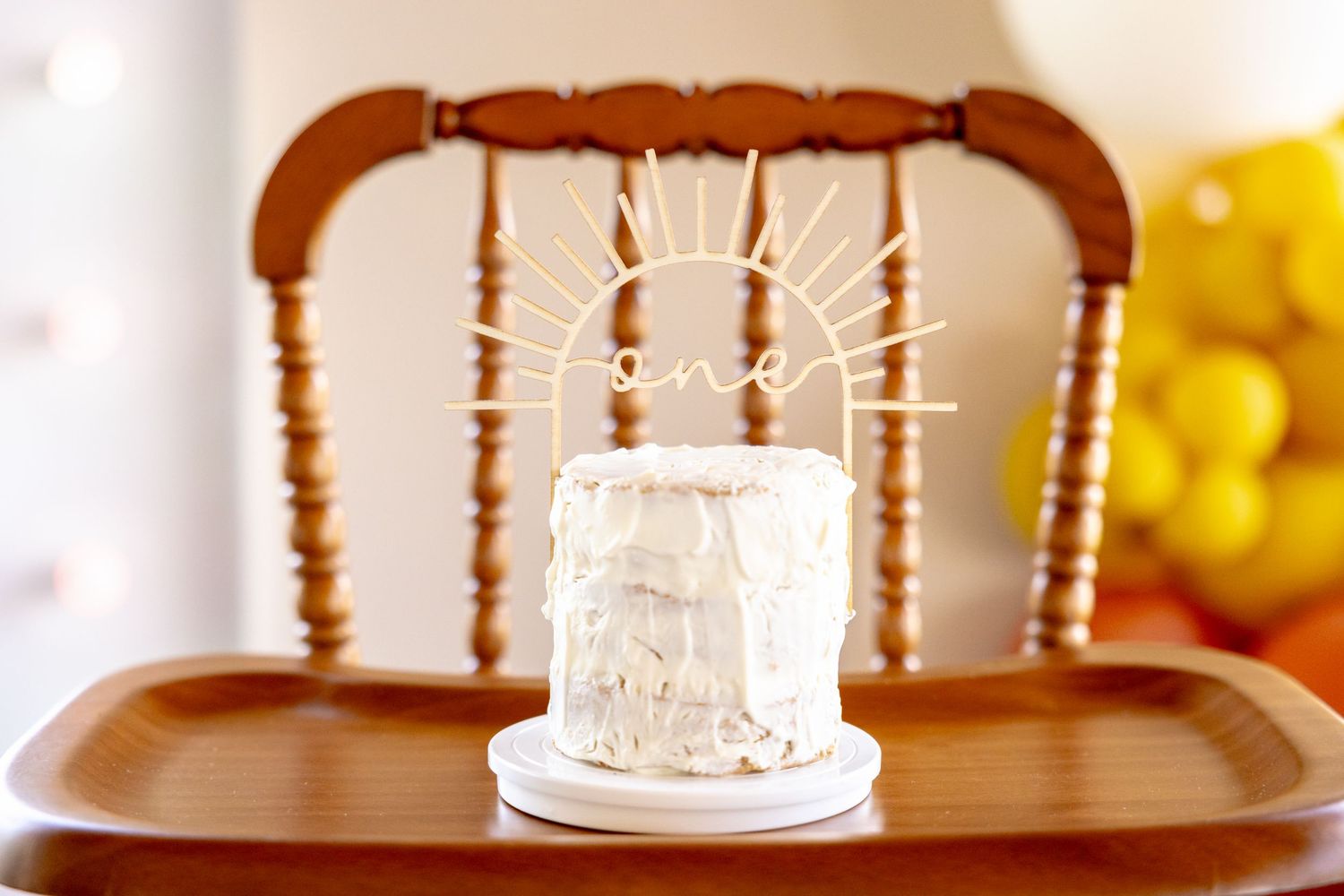 White smash cake sitting on wooden vintage high chair with neutral party decorations.