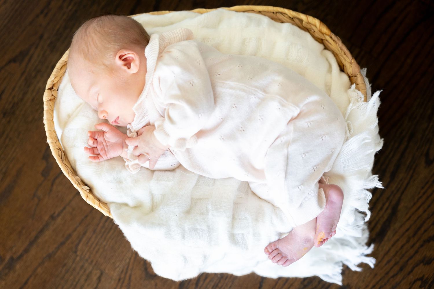 Baby wearing white ruffled dress resting in woven basket on dark wooden floor.