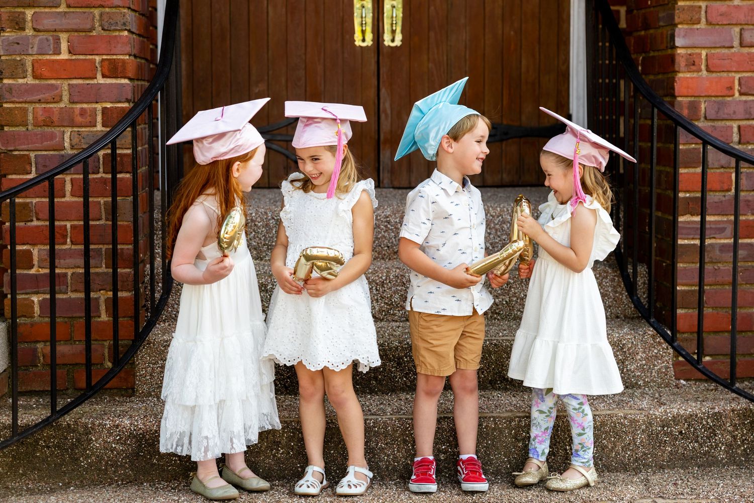 Group of young children in preschool graduation attire lined up against brick wall and wooden doors.