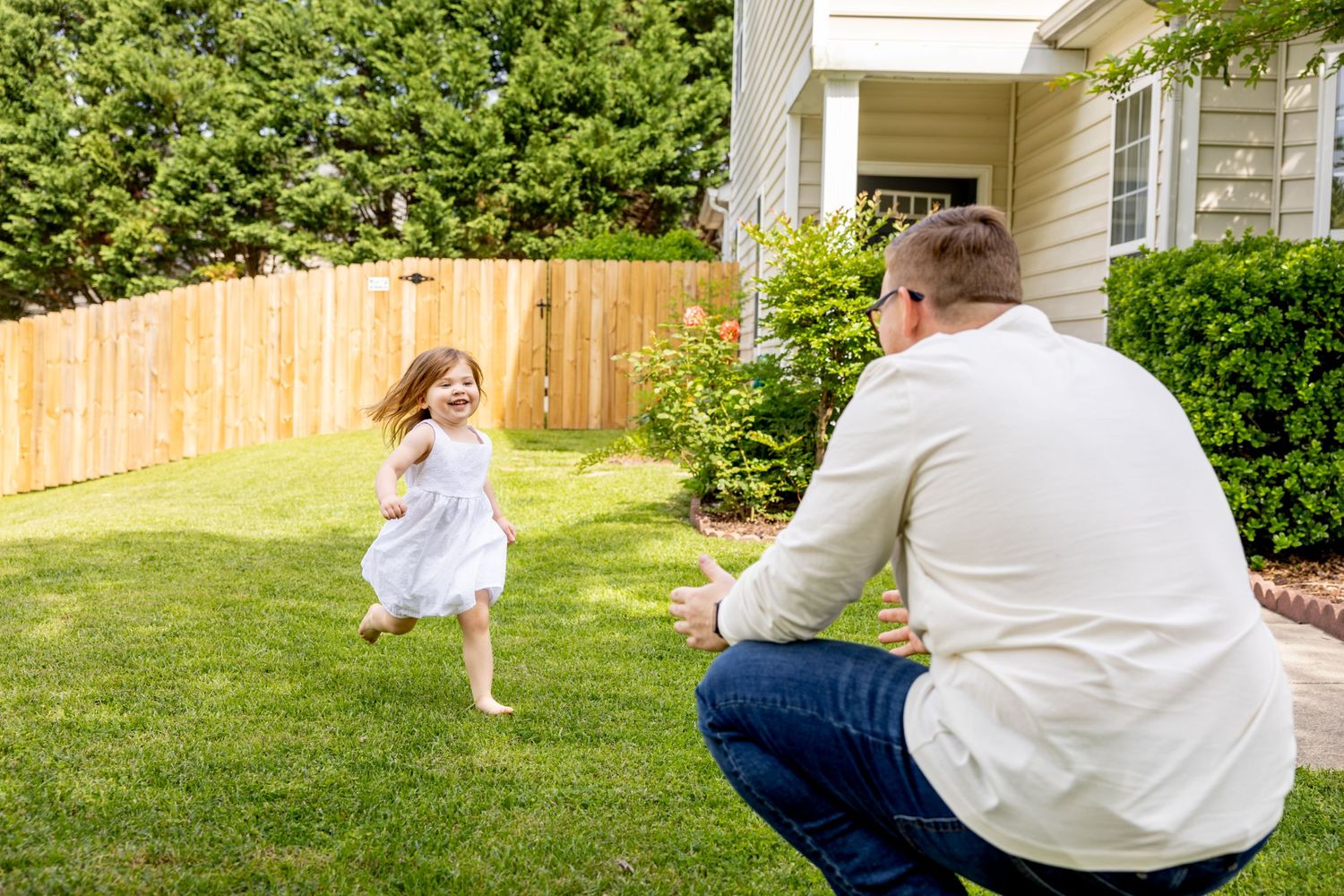 Child in white dress playing in backyard while adult watches from porch.