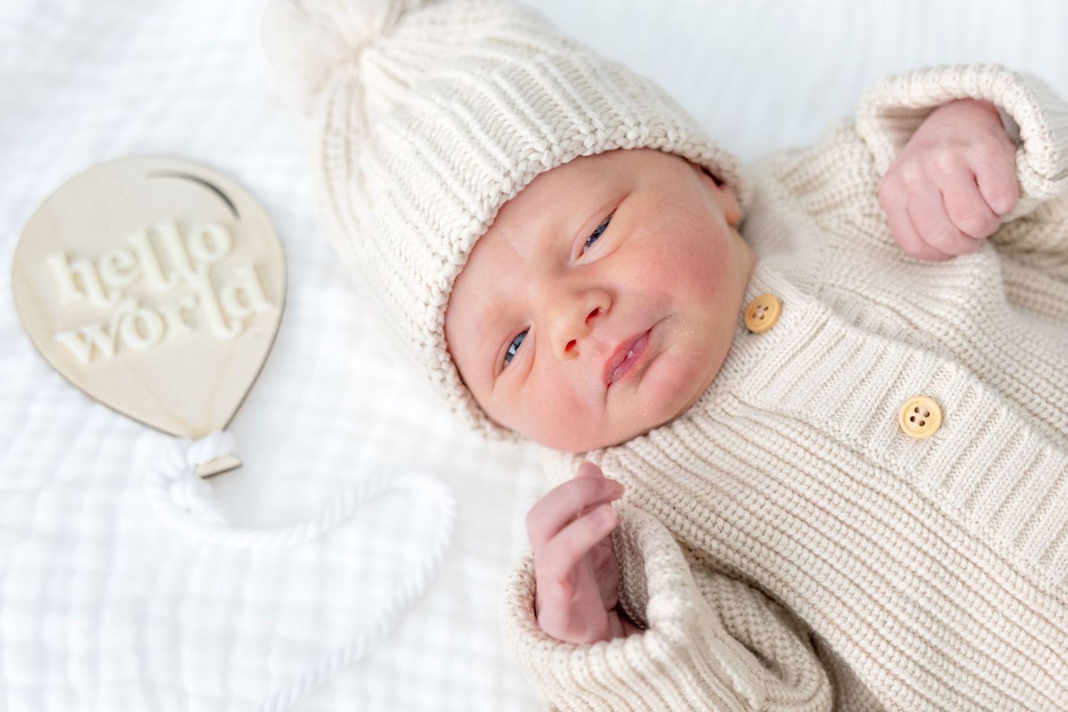 Newborn in white knit outfit lying next to 'hello world' sign on white bedding.