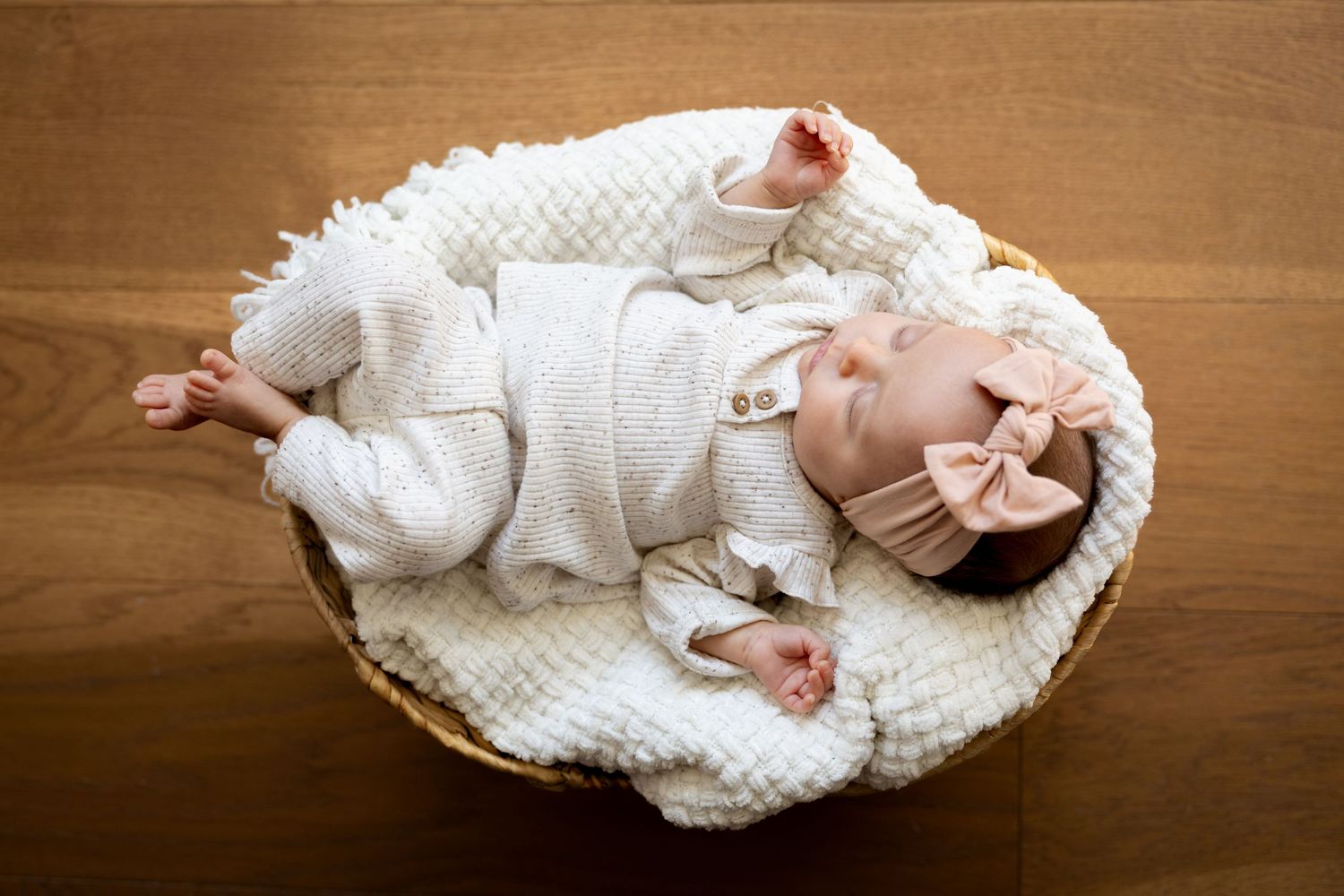 Overhead view of sleeping baby in white outfit lying in woven basket on wooden surface.