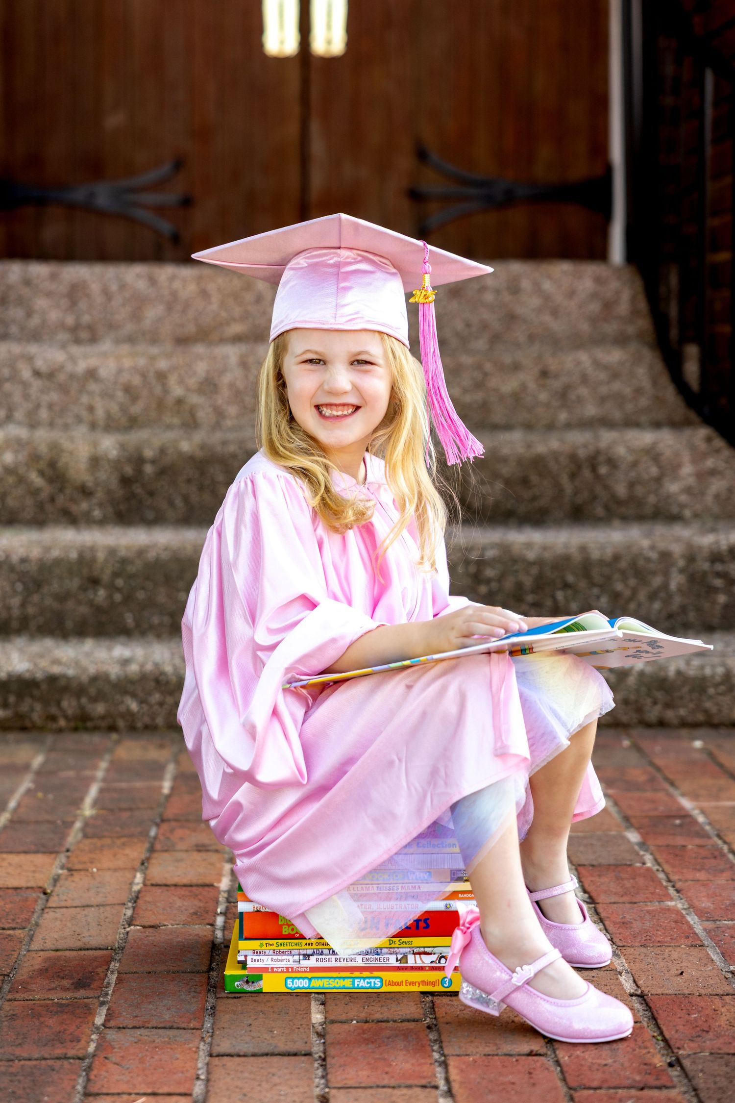 Graduate in pink sits on stack of books on brick steps with book in hand.