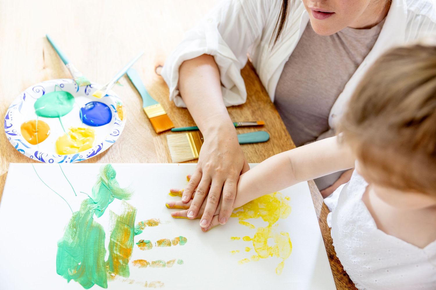 Hands create colorful artwork together using yellow and green paint on white paper.
