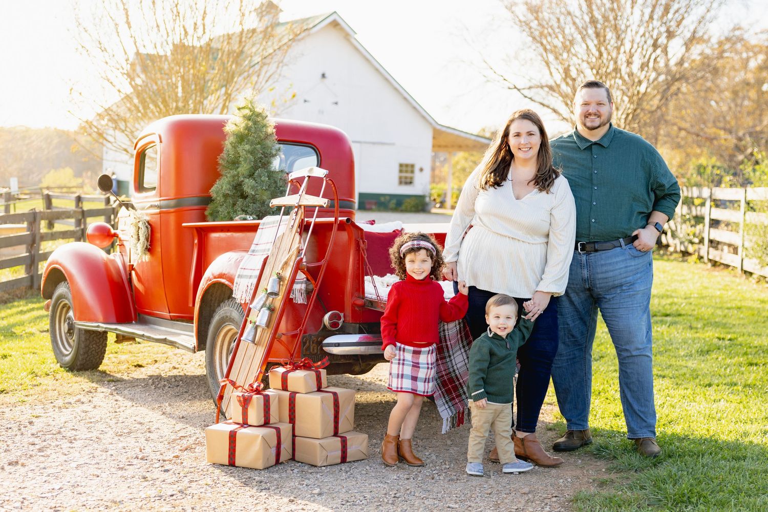 Family poses with vintage red truck and wrapped presents in rustic farm setting.
