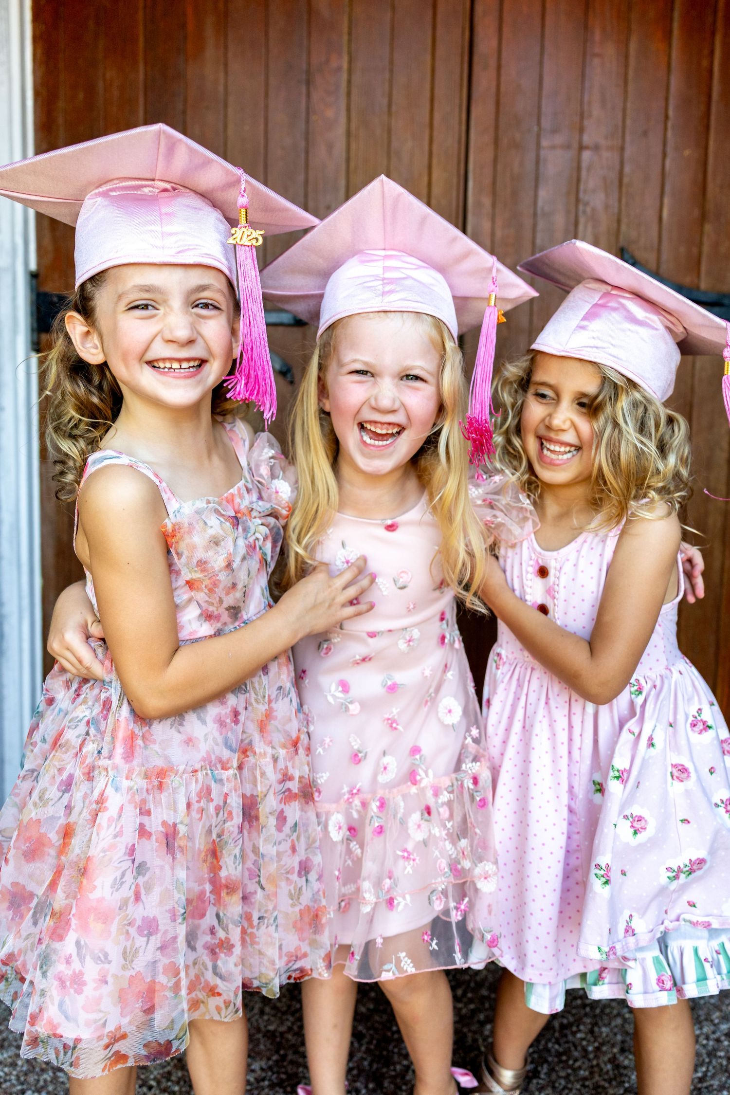 Group of preschool graduates in floral dresses and pink graduation caps laughing together.
