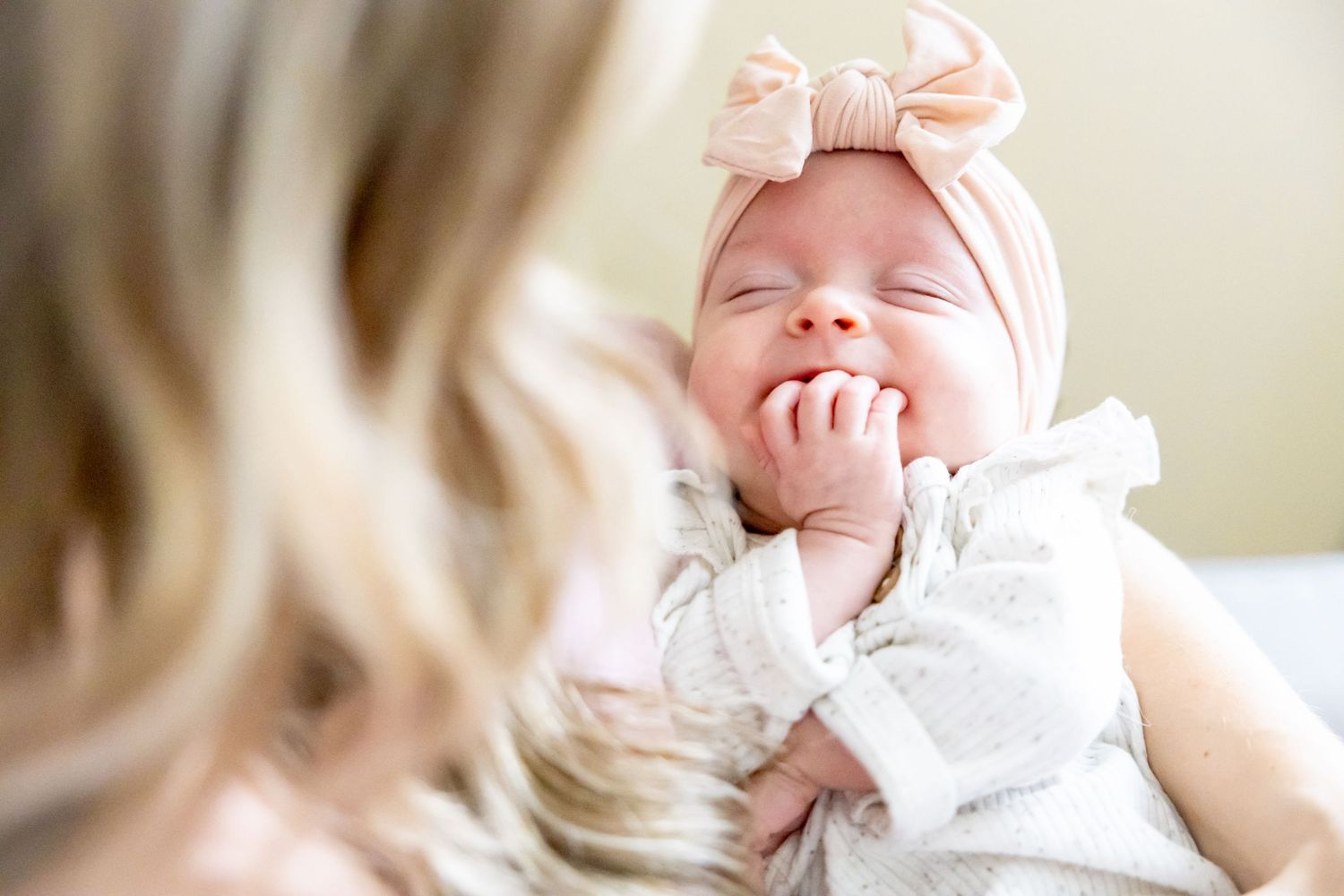 Baby wearing white outfit and pink bow headband smiles while being held.