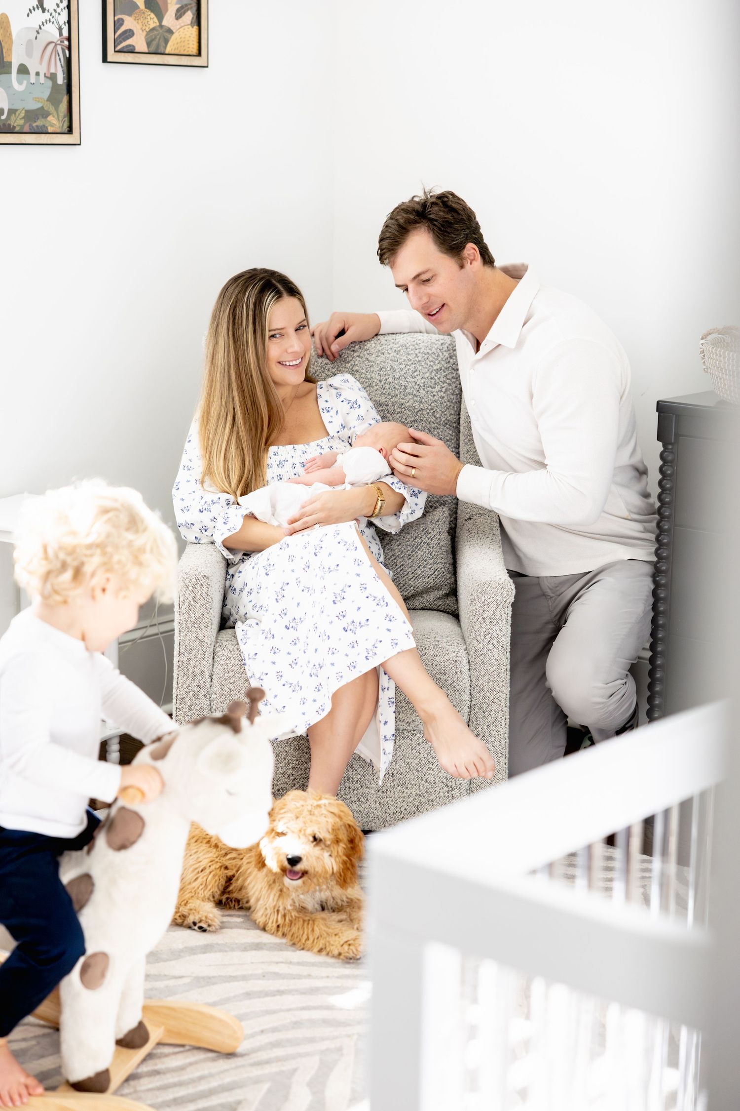 Family shares tender moment on grey armchair while dog rests nearby and toddler plays.