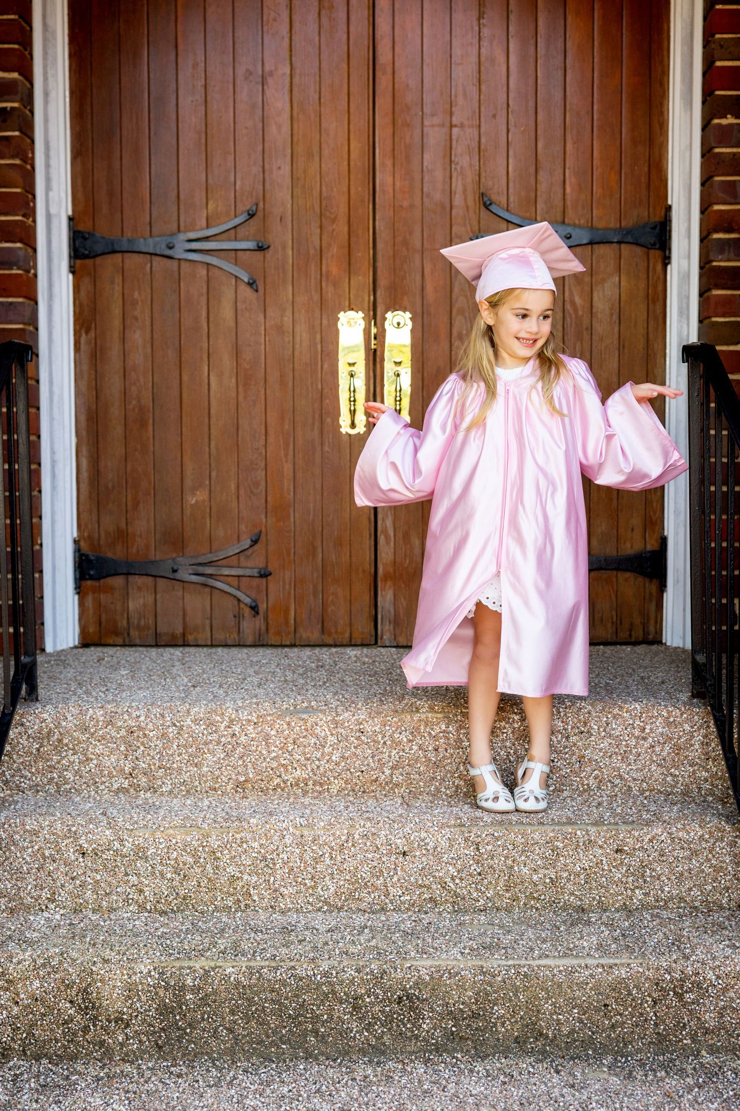 Child in pink preschool graduation attire posing in front of wooden church doors.
