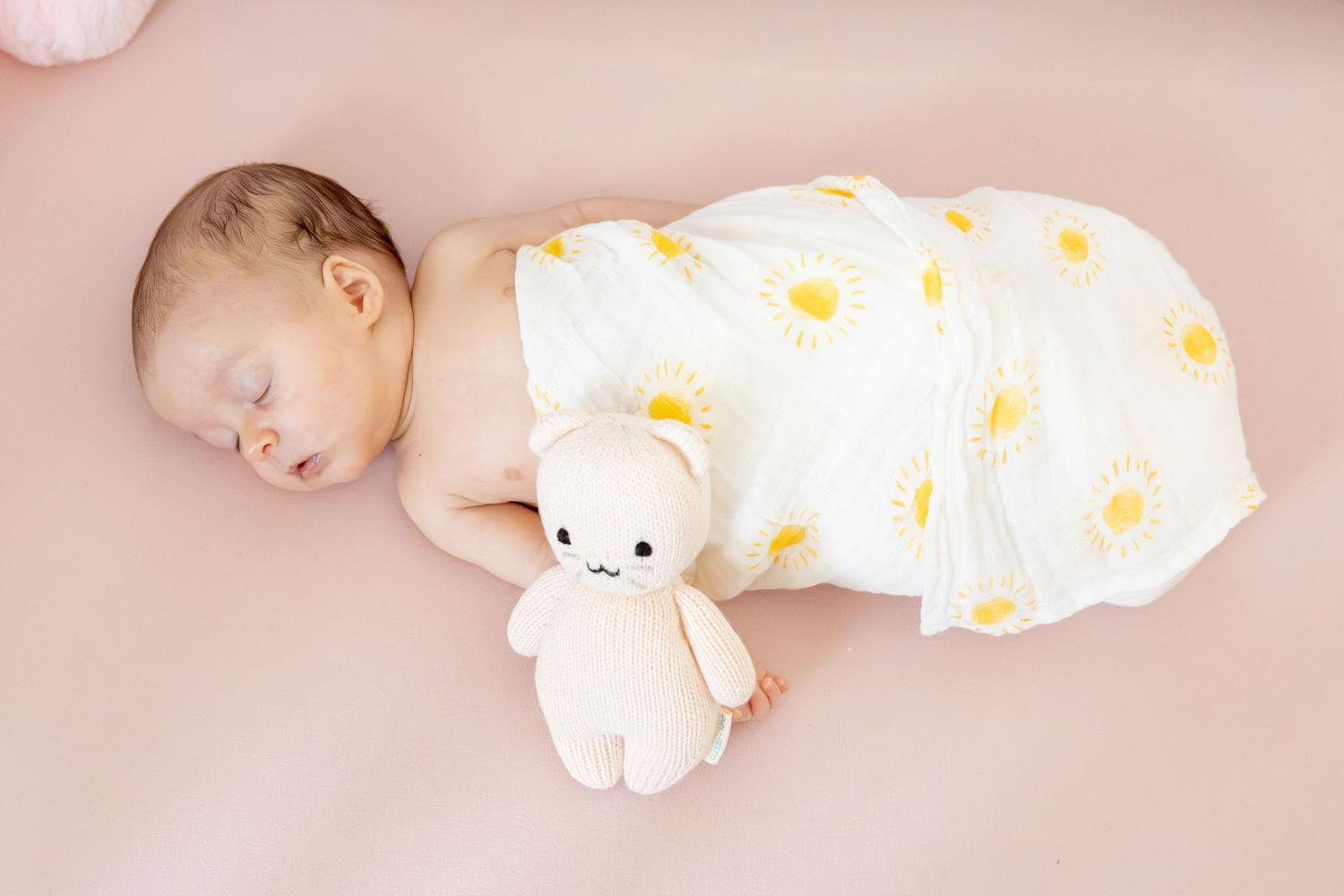 A sleeping baby in a polka dot outfit holding a white stuffed toy on pink bedding.
