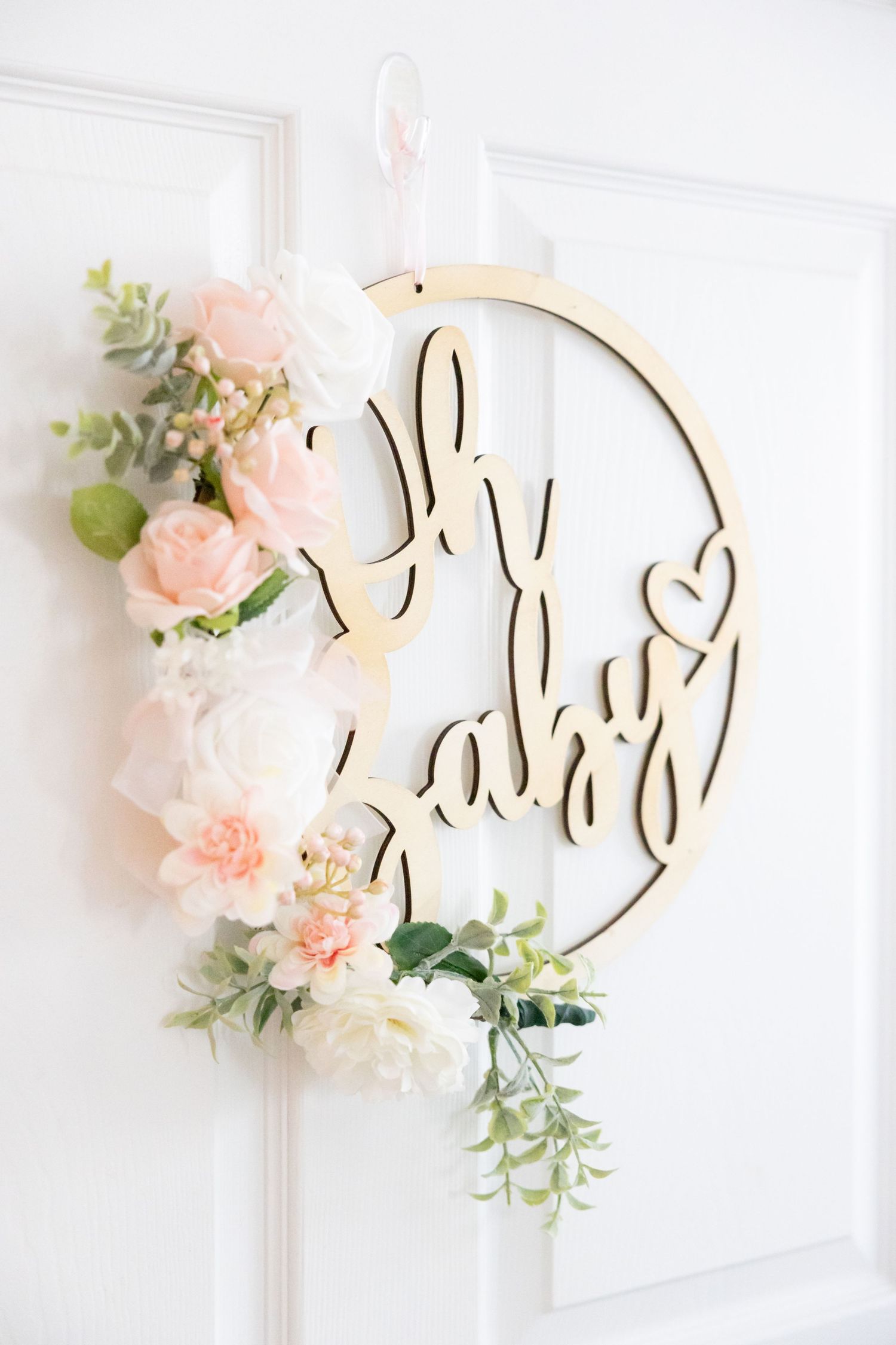 Decorative wooden 'Oh Baby' wreath with pink and white flowers hanging on white door.