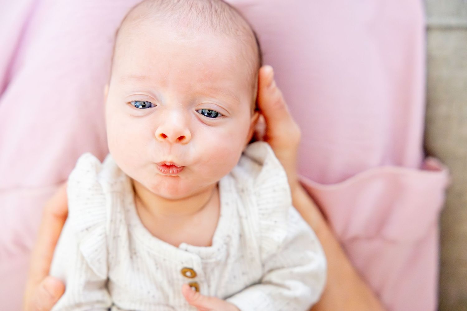 Newborn baby in white outfit makes adorable expression against pink background.