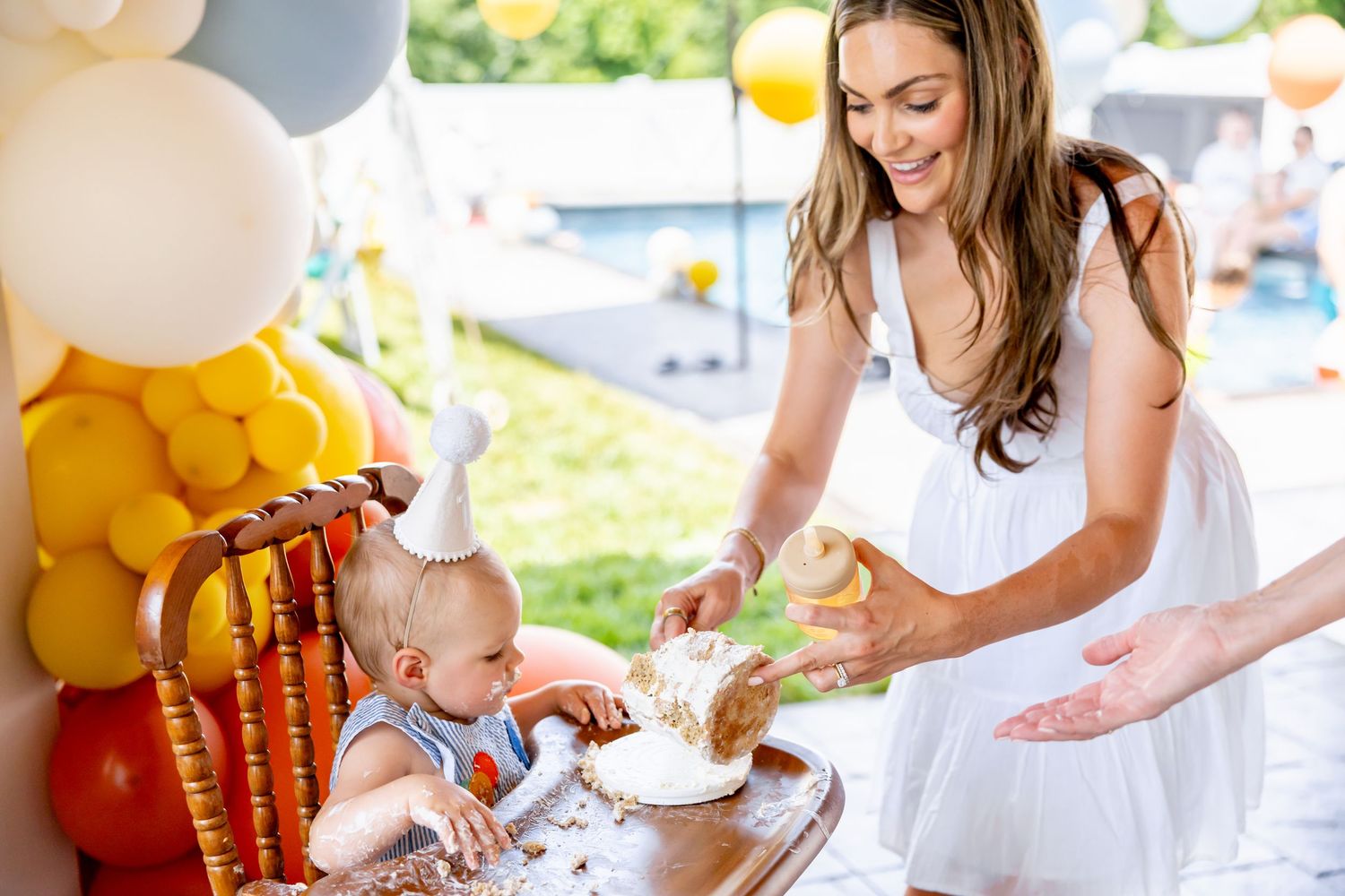 Birthday party scene with orange and yellow balloon decorations and white-dressed attendees.