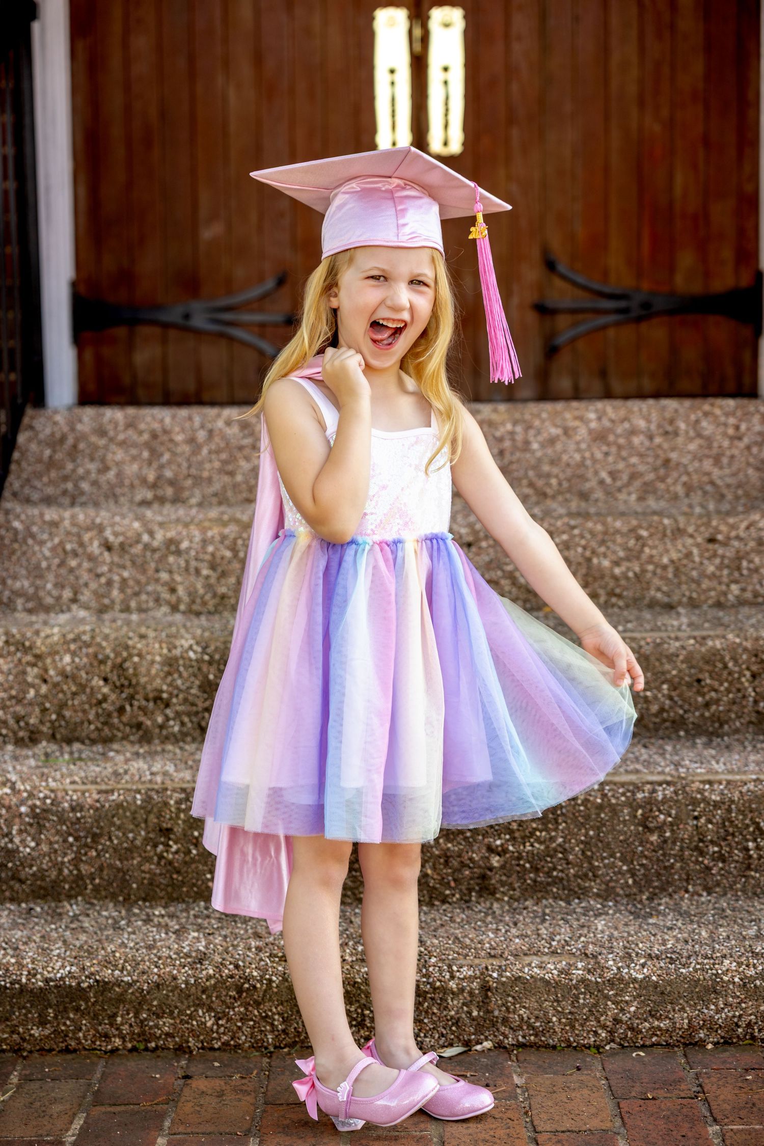 A small kindergarten graduate in a pink and white frilly dress and shoes standing on a brick walkway.