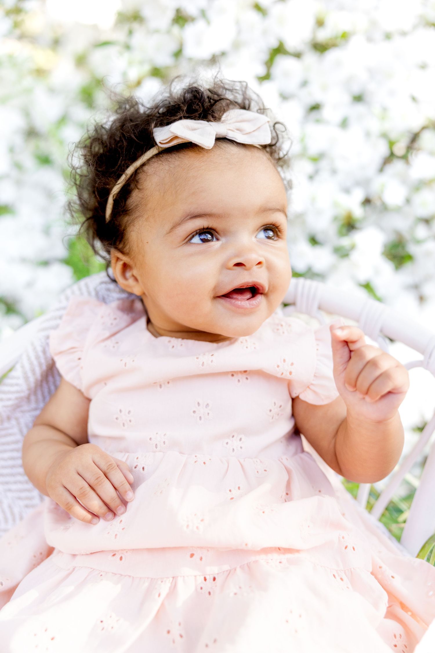 Baby wearing pink dress and white bow headband sitting among spring flowers at Glencairn Gardens in Rock Hill, SC.