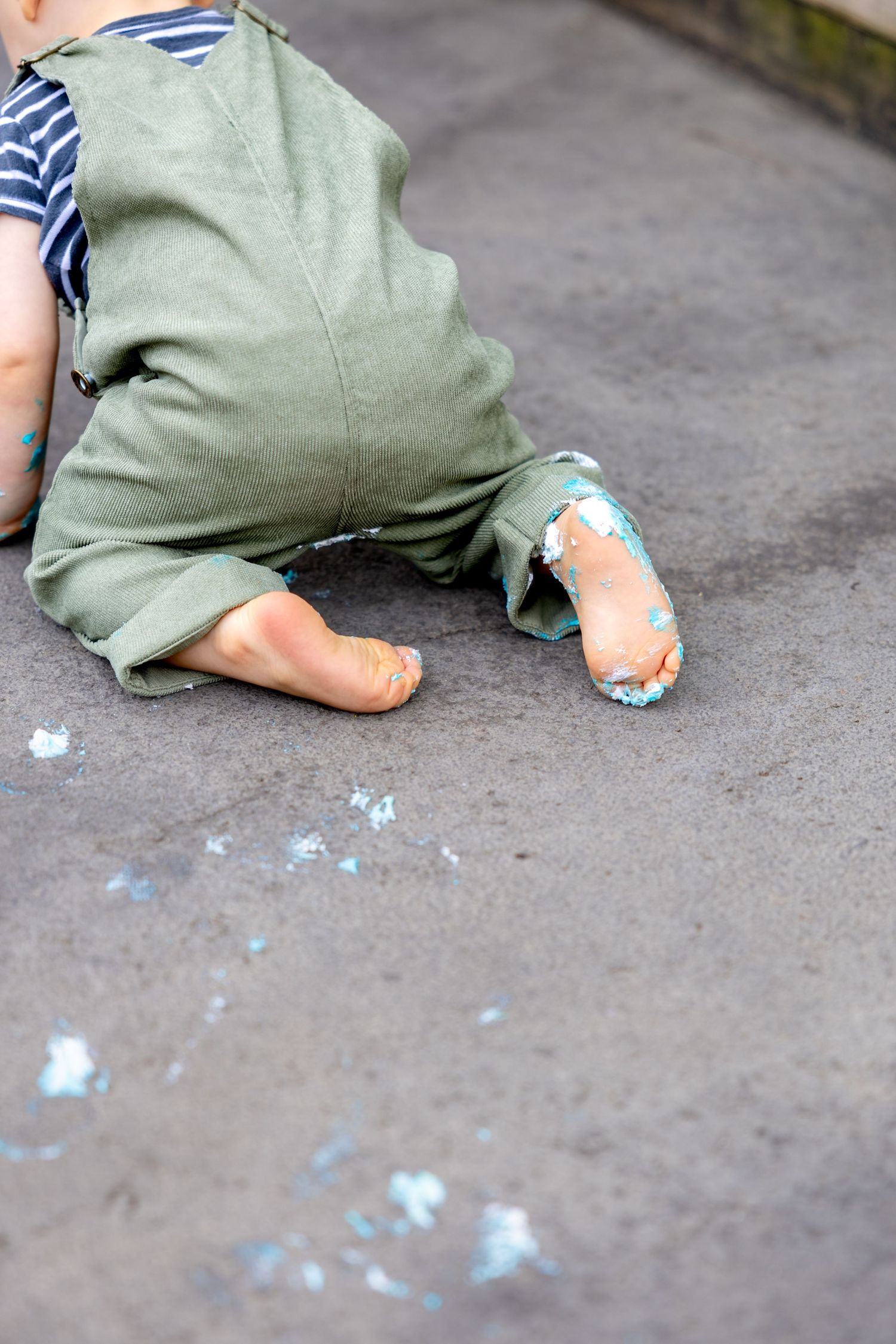 Little feet in green overalls with blue frosting marks on concrete ground after successful cake smash session.