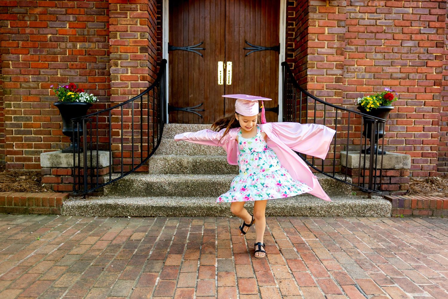 Pink-clad preschool graduate twirls joyfully on brick steps in front of wooden doors.